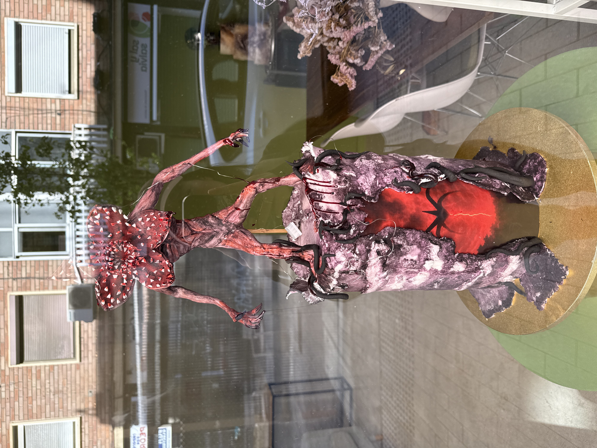 The image depicts a diorama of the Demogorgon character from the "Stranger Things" television series, displayed behind a window. The Demogorgon figure is predominantly red and textured, with its characteristic floral head. The diorama is set on a gold circular base, and includes elements of a dark, menacing environment, possibly representing the Upside Down. The window's reflection shows a brick building with windows, and a sign with "Divide" is visible, suggesting a commercial or service establishment.
Based on the sign "Divide," it is possible that this image was taken in Barcelona, Spain, given that "Divide" is a common name for companies in the area. The image's location in Barcelona cannot be completely verified without further information.