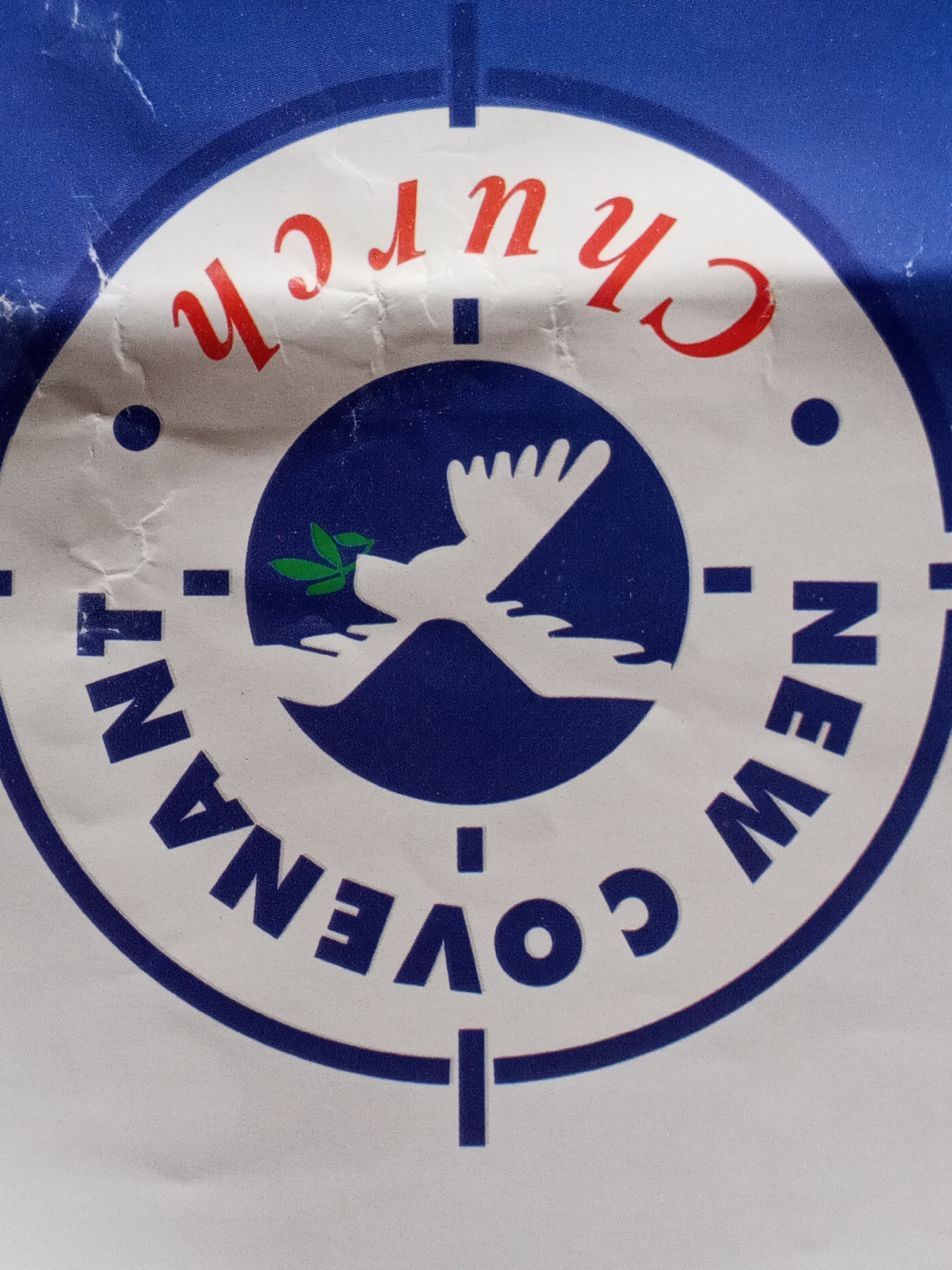 The image presents a close-up of a circular logo, likely for a religious institution. The design is set against a background that transitions from solid blue at the top to white within the logo's main boundary.
At the core of the logo is a stylized depiction of a white dove in flight, carrying a vibrant green olive branch in its beak, all set against a dark blue circular backdrop. Beneath the dove, two white hands are rendered, appearing to gently support or cradle the bird.
Encircling this central emblem, within a wider white band, is text. The words "NEW COVENANT" are visible in dark blue, capital letters, positioned at the bottom of the circle. At the top of the circle, oriented upside down, the word "CHURCH" is displayed in red, capital letters. The overall circular logo is outlined by a thin dark blue border, which also includes dashed lines and dots at various points, reminiscent of a target or compass.
The media appears to be a printed document, showing signs of wear such as creases and minor surface imperfections, indicating it has been handled. The colors are distinct and bold. This logo is associated with a church in Badagry, Nigeria.