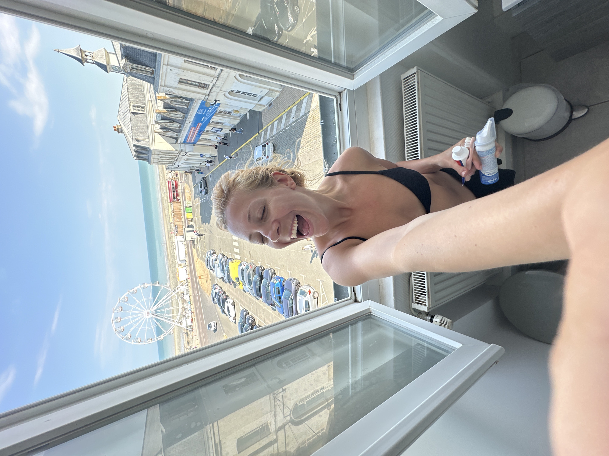 In Le Tréport, France, a woman with blonde hair tied back is positioned in the foreground, leaning out of an open window and taking a selfie. She is wearing a black bikini top and black bottoms. Her mouth is open in a wide smile, and she is holding a tube of toothpaste and a toothbrush in her right hand. The background reveals a street scene with parked cars, buildings, and the sea with a Ferris wheel in the distance. The overall environment suggests a sunny day.