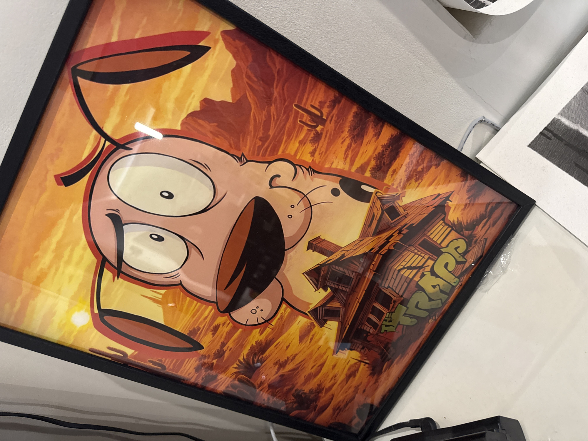 A framed poster of Courage the Cowardly Dog is displayed on a white surface, leaning against a white wall. The poster depicts Courage in a desert landscape with a rundown house in the foreground. The house has graffiti on it that reads "THE TRAP". A black cable is visible on the left side of the image, running down the wall. Part of another piece of artwork is visible in the lower right corner of the image. The overall scene suggests an indoor setting, possibly a home or an office, with no specific identifiable landmarks of Sofia, Bulgaria.