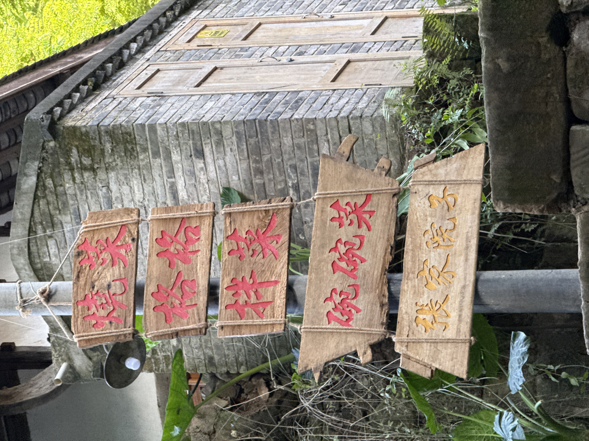 The image depicts a street scene in Beibei, China. Five weathered wooden signs with carved Chinese characters hang from a metal bar. The characters on the first four signs are painted red, while the characters on the fifth sign are painted gold. Behind the signs is a traditional Chinese building with a grey brick facade and a wooden roof. Vegetation grows on the building and in the surrounding area. A lamp is affixed to the metal bar.