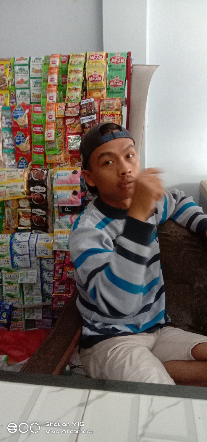 A person wearing a blue-striped sweater and a baseball cap sits in front of a wall lined with rows of packaged snacks, including items like "MP-ASI," "Desaku," "Ladaku," "WOOMILK," "posh," and "chocolatos." The individual has a hand near their face, perhaps in a gesture. A camera watermark is visible at the bottom of the image, indicating it was taken with a Vivo Y15. The setting appears to be a small shop or kiosk.