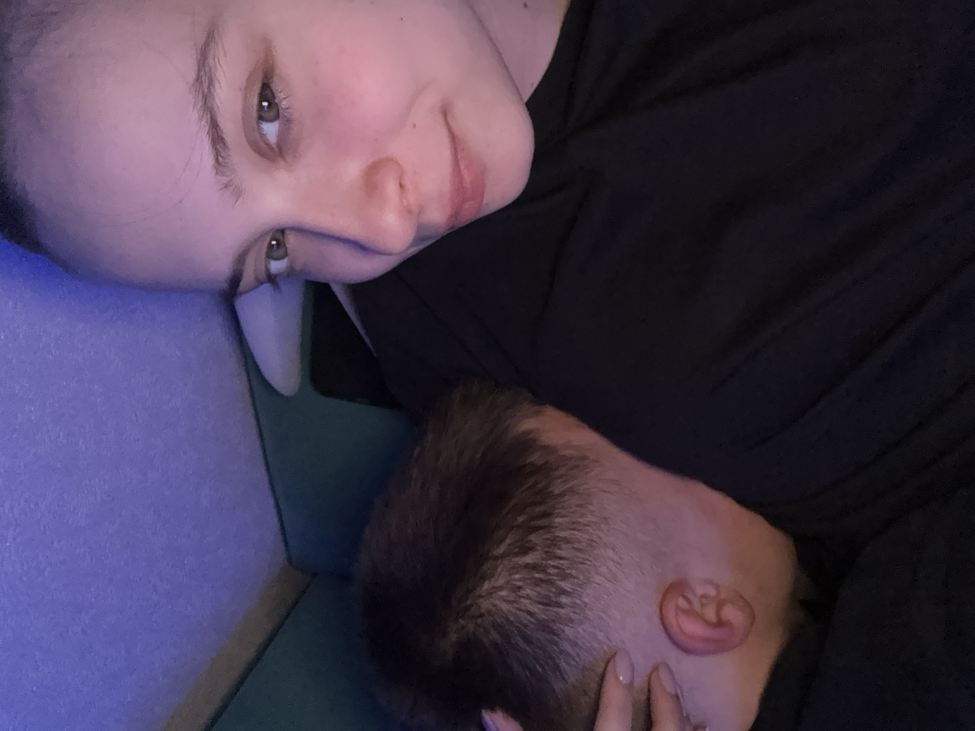 A selfie-style image shows a woman lying down with a man's head resting on her. The woman has fair skin, light brown eyes, and light eyebrows. She wears a dark-colored shirt. The man has short, dark hair. The setting appears to be indoors, possibly on a couch or bed, with a blue wall visible in the background. There is no explicit information to confirm the location in Прикамский, Russia.