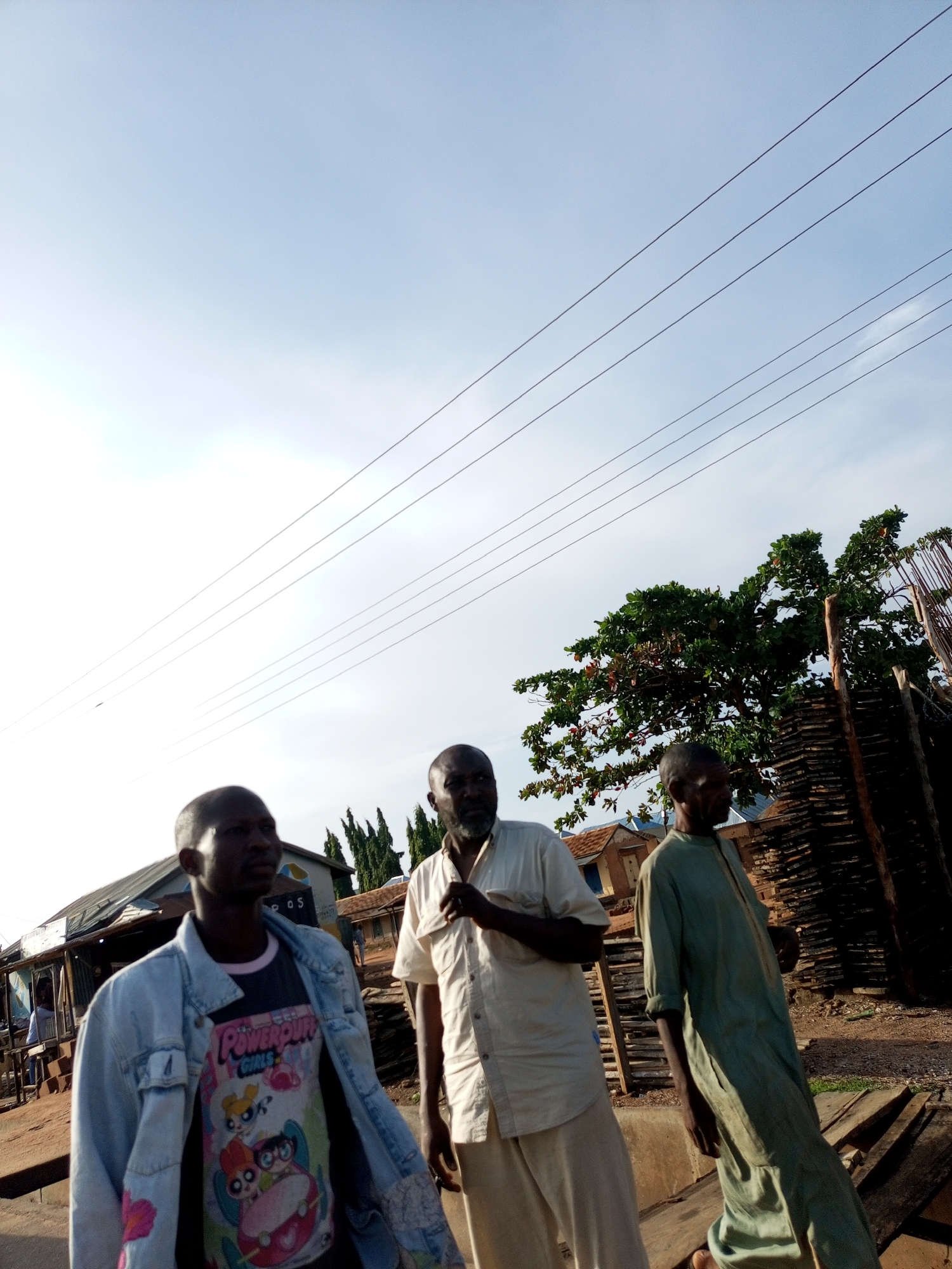 Three men are standing outdoors in what appears to be a marketplace or roadside area in Tumfure, Nigeria. The man on the left is wearing a light blue denim jacket over a t-shirt with a Powerpuff Girls graphic. The middle man is wearing a cream-colored shirt and pants. The man on the right is wearing a long, light green robe. In the background, there are wooden structures, a tree with green foliage, and power lines stretching across the sky. The sky is clear and bright.