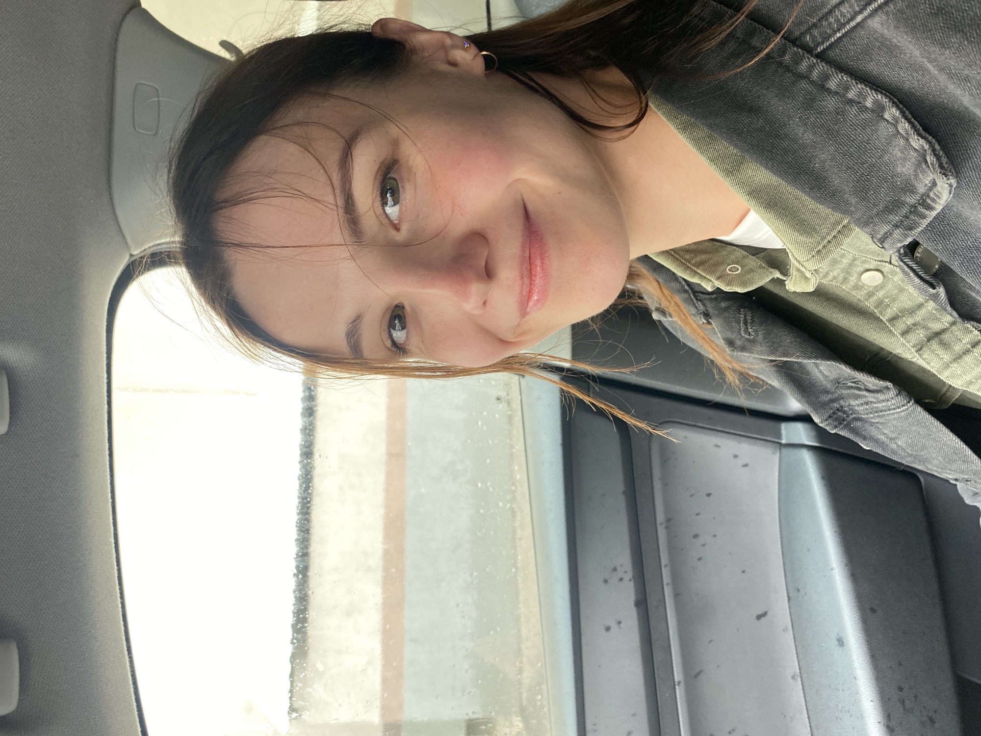A young woman with brown hair and green eyes smiles at the camera from the passenger seat of a car. She is wearing a green collared shirt under a dark denim jacket. The car's interior is visible, including the door panel and the window. Outside the window, it appears to be raining, with water droplets on the glass and a blurry background suggesting an urban environment. The image was taken in Novokuybyshevsk, Russia.