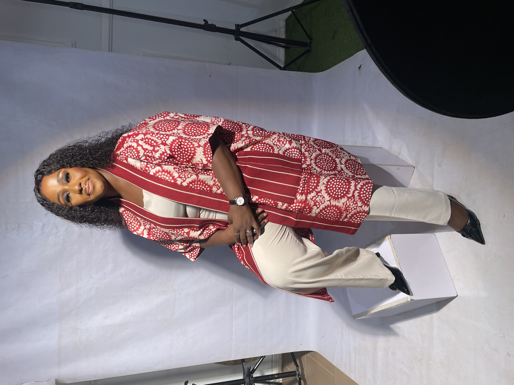A woman poses indoors on white steps against a white backdrop. A light stand and diffused light source are visible. She wears a cream-colored top and pants with black shoes. A patterned red and white shawl drapes around her shoulders. She has dark curly hair and is wearing a watch and rings. The location is identified as Jos, Nigeria.