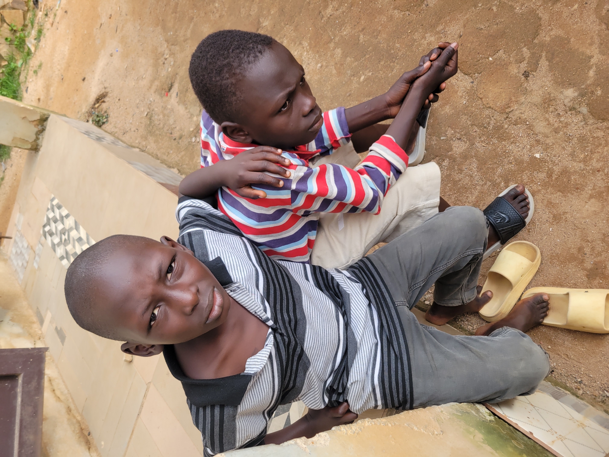 Two young Black boys are pictured outdoors, sitting close together. The boy in the foreground is looking directly at the camera with a neutral expression, wearing a black and grey striped shirt and grey pants. He has short, dark hair. The second boy is sitting behind him, looking upwards and to his left. He is wearing a striped shirt with red, blue, and white stripes, and is holding the first boy's hands. Both boys are barefoot, with one boy wearing a black sandal on his foot. They are sitting on a textured, dirt-like surface next to a light-colored tiled area. In the background, there is more dirt and some green vegetation. The lighting suggests it is daytime. The scene appears to be in Jos, Nigeria.