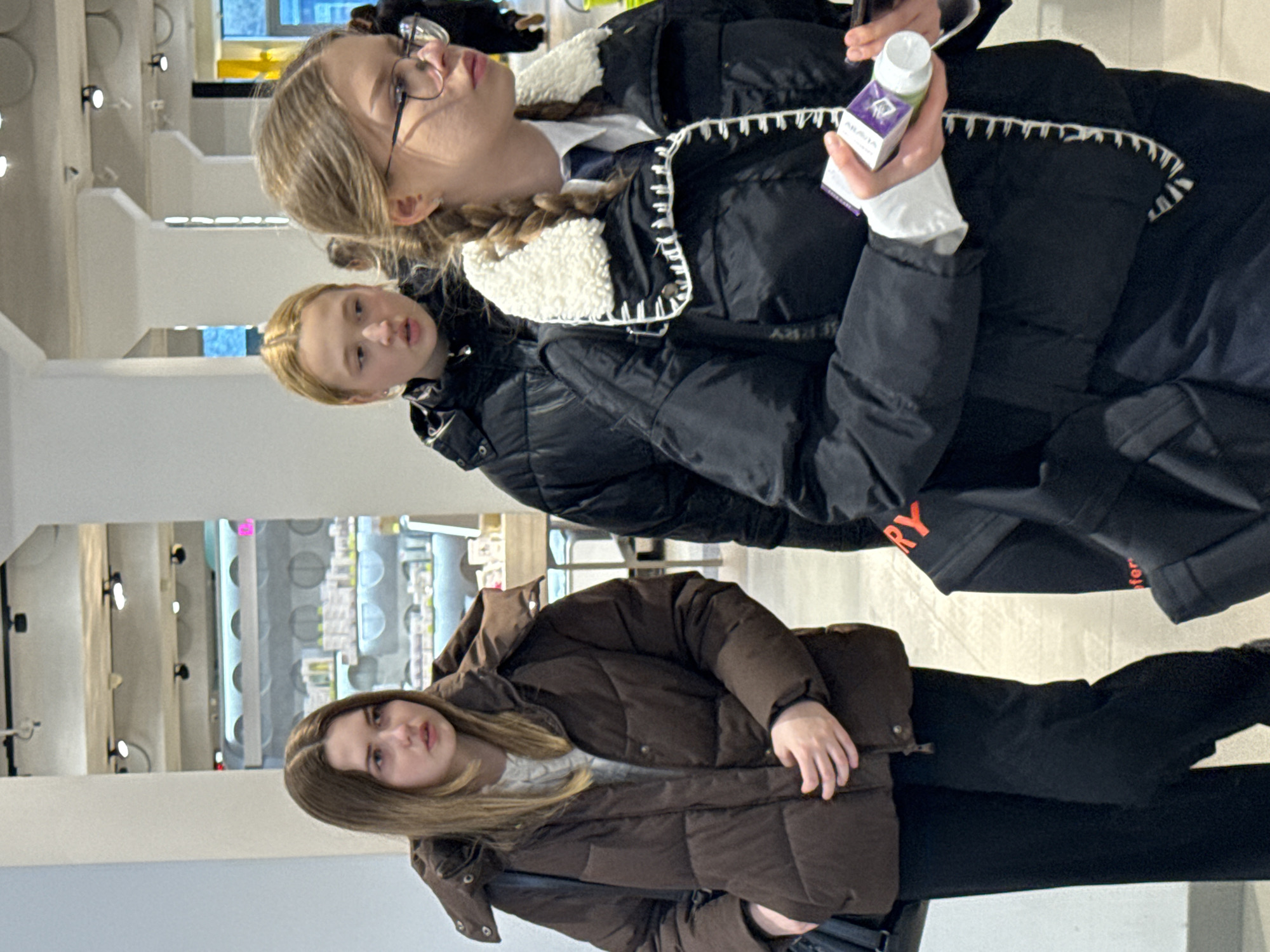 Three young women are standing in a store in Kazan, Russia. The store appears to sell skincare or cosmetic products. The first woman, in a brown puffer jacket, stands at the bottom of the image. Above her stands another young woman in a black puffer jacket. The third woman, in a similar black puffer jacket with a white collar, is holding a small, purple and white box. She is wearing glasses and her hair is in braids. The store has white walls and shelving. There is bright lighting in the store.