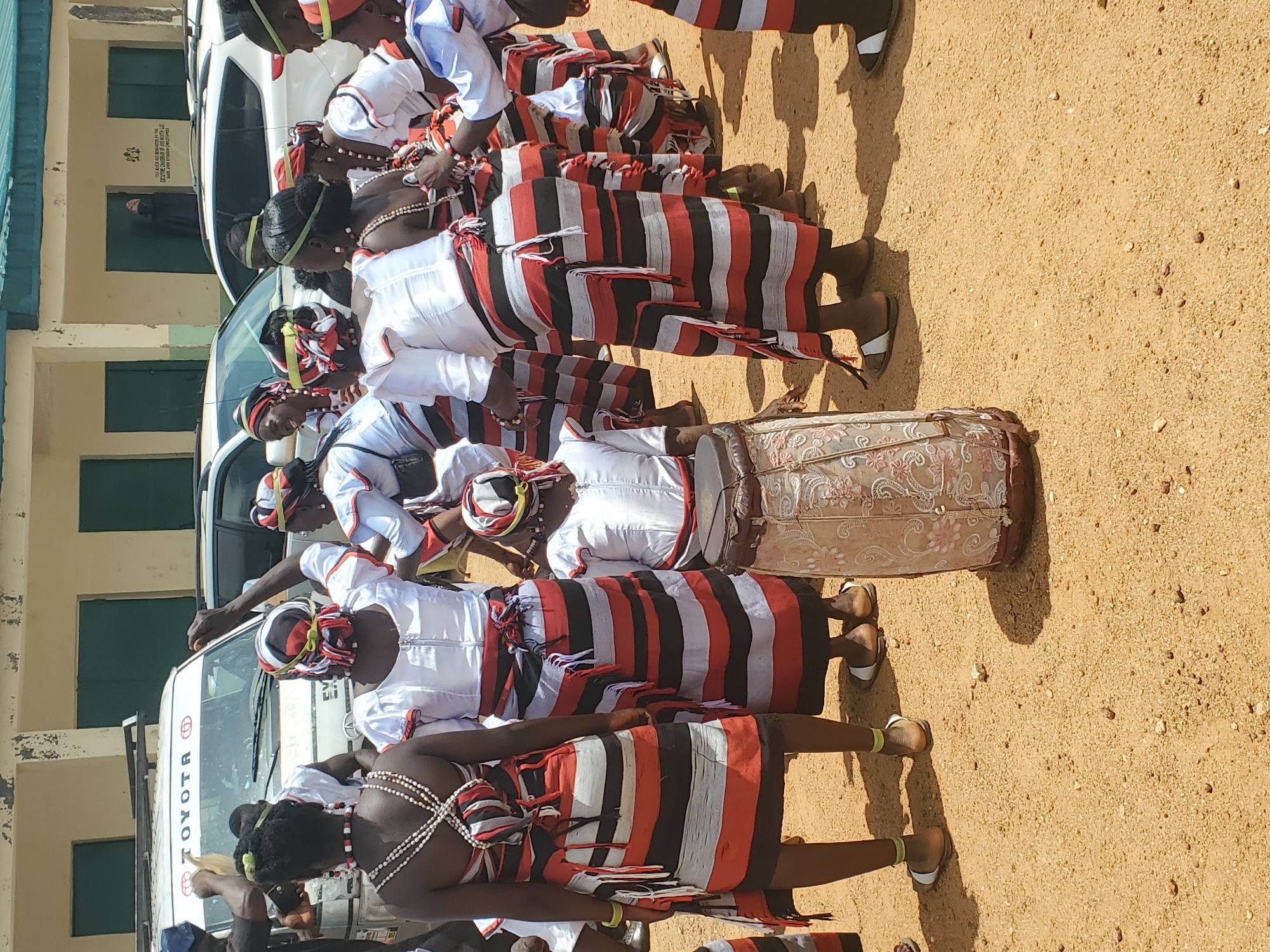 This is a medium shot, taken at eye level, of a group of people in traditional attire, gathered outdoors in a dusty, sandy area. The setting appears to be a village or rural area within Kabong, Nigeria, suggested by the presence of a building in the background with visible green trim and a sign that reads "TOYOTA" on a white van. Several individuals are dressed in matching white tops and brightly colored striped skirts made of red, black, and white fabric. Many are also wearing headbands in similar colors. One person in the foreground, facing away from the camera, is playing a drum with an intricately patterned design. The people are interacting, some appearing to be dancing or engaged in a communal activity. The lighting indicates it is daytime, with shadows cast on the ground, suggesting a sunny environment.