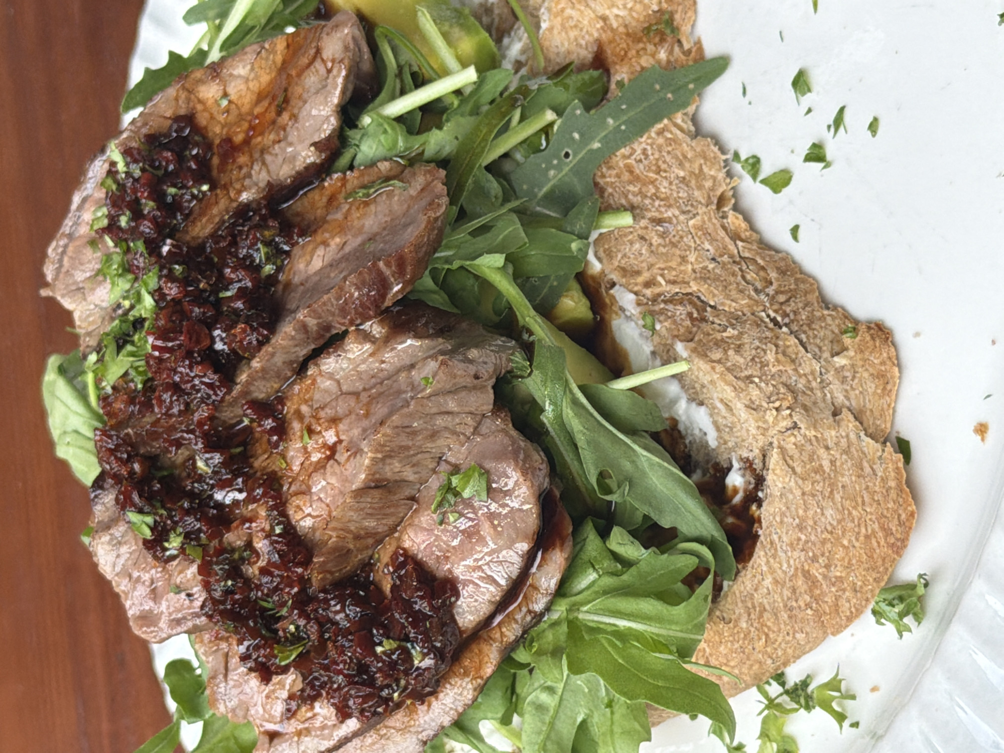 The image provides an overhead, close-up view of a meticulously prepared meal presented on a white plate. The central components of the dish are several slices of seared, cooked meat, likely beef steak, which exhibit a tender, medium-rare appearance in some areas. These meat slices are generously topped with a dark, reddish-brown relish or compote, possibly made from finely chopped dried fruits like cranberries or dates, or caramelized onions, creating a rich textural contrast. Freshly chopped green herbs, most likely parsley or cilantro, are sprinkled over both the meat and the relish, adding a touch of freshness and color.
The sliced meat rests on a vibrant bed of fresh green leaves, primarily identified as arugula due to its distinctive shape and size. Interspersed among the greens and underneath the meat, thin slices of avocado are visible, contributing another layer of color and richness to the assembly. Adjacent to the meat and greens, and partially covered by them, are several pieces of light brown, textured flatbread or pita bread, appearing somewhat rustic with a slightly crumbly or crispy surface. A creamy white substance, possibly a yogurt-based sauce, labneh, or a light dressing, is spread beneath some of the flatbread and other ingredients, providing a moist base.
The white plate serves as a clean backdrop for the colorful and varied components of the meal. A dark wooden surface, possibly a tabletop, is visible in the upper and lower left corners of the frame, hinting at the setting. The image is solely focused on the food, capturing its textures and presentation in detail. There are no people visible, nor any discernible text. The dish appears to be a savory, wholesome meal, likely enjoyed for lunch or dinner, presented in a style that could be found in a contemporary restaurant or café, possibly within the context of Đà Nẵng, Vietnam.