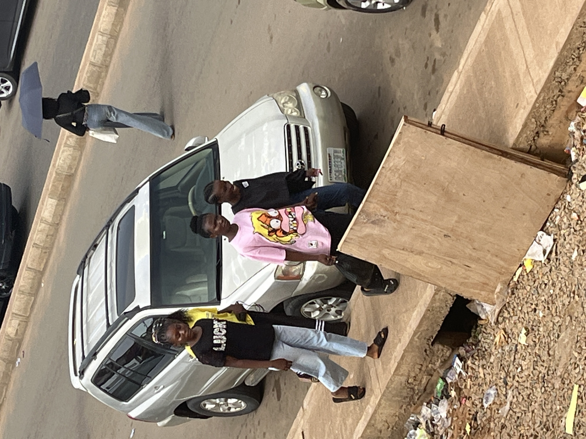 An overhead, eye-level, medium shot captures three young women walking on a sidewalk next to a silver SUV on a street in Jos, Nigeria. The woman in the foreground on the left wears a black t-shirt with the word "LUCKY" in white lettering, and light blue jeans. The middle woman wears a pink t-shirt with a cartoon character and dark blue jeans, and carries a black bag. The woman on the right wears a black long-sleeved shirt and dark blue jeans. The sidewalk is lined with rubble, debris, and a wooden plank covering a drainage ditch. In the background, a woman holding a dark blue umbrella walks away from the camera. The scene appears to be a typical streetscape in Jos, Nigeria, characterized by its urban environment and pedestrian activity.