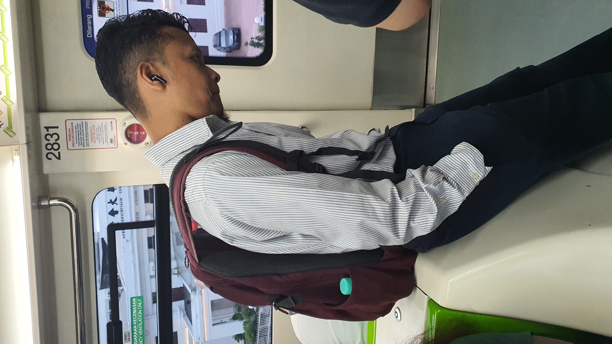 This media file captures a medium shot of a man sitting inside a train, likely in Kuala Lumpur, Malaysia, given the text present. The man is wearing a light blue and white striped dress shirt, dark trousers, and has a maroon backpack slung over his shoulders. He also appears to be wearing wireless earbuds. His legs are crossed, and he is leaning back against the seat. The overall impression is of a commuter traveling.
The setting is clearly a train carriage, indicated by the seats, windows showing passing scenery (including a car outside), and various labels and signage on the train's interior. One prominent sign displays the number "2831" and text in Malay, including "PEMBUKA PINTU KECEMASAN" which translates to "EMERGENCY DOOR RELEASE." Another sign states "DILARANG PRO" (PROHIBITED). Other visible text includes "MR11," "MR10," and "MR9," which could refer to specific train lines or carriage numbers. There are also some Chinese characters visible through the window, suggesting a culturally diverse area.
The lighting suggests it might be daytime, but without further information, the specific time of day and weather conditions cannot be determined. The man's facial expression is neutral, not conveying any strong emotions. The activity is passive; he is simply a passenger traveling. The image is oriented vertically, implying it was taken with the camera held in portrait mode.