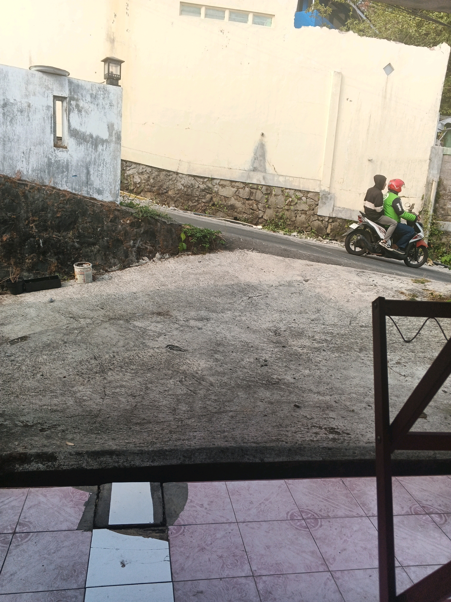 In Keji, Indonesia, a motorcycle carrying two individuals travels uphill on a paved road. The rider, wearing a red helmet, is dressed in a dark hoodie with white accents. The passenger, in a bright green jacket, also wears a dark hoodie. They are moving away from the viewer towards a pale yellow building with a rough, stone-based retaining wall. The road is bordered by this wall and a concrete area in the foreground. To the left of the road, a rough-textured, pale grey wall with a small, rectangular opening and a black outdoor light fixture stands against the backdrop of the pale yellow building. In the foreground, a cracked concrete surface transitions to a tiled patio with a dark metal structure partially obscuring the view. The scene is lit by natural daylight, casting shadows.