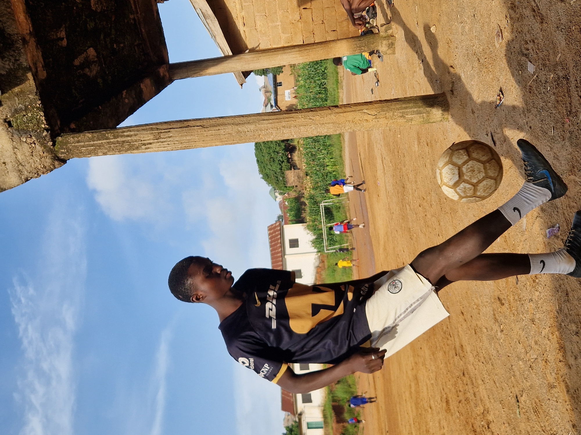 The image depicts a young Black male playing soccer on a dirt field in Kabong, Nigeria. He is wearing a dark blue and gold soccer jersey, light-colored shorts, white socks with a Nike logo, and dark cleats. The man is in mid-action, with his right foot extended to kick a soccer ball. The soccer ball is partially in the air, to the right of the player. In the background, other individuals can be seen playing soccer, with a goalpost visible. The environment includes sandy dirt ground, some vegetation, and buildings in the distance, suggesting an urban or semi-urban setting. The scene is partially shaded by an unfinished structure with thick, weathered concrete pillars. The sky is blue with scattered clouds.