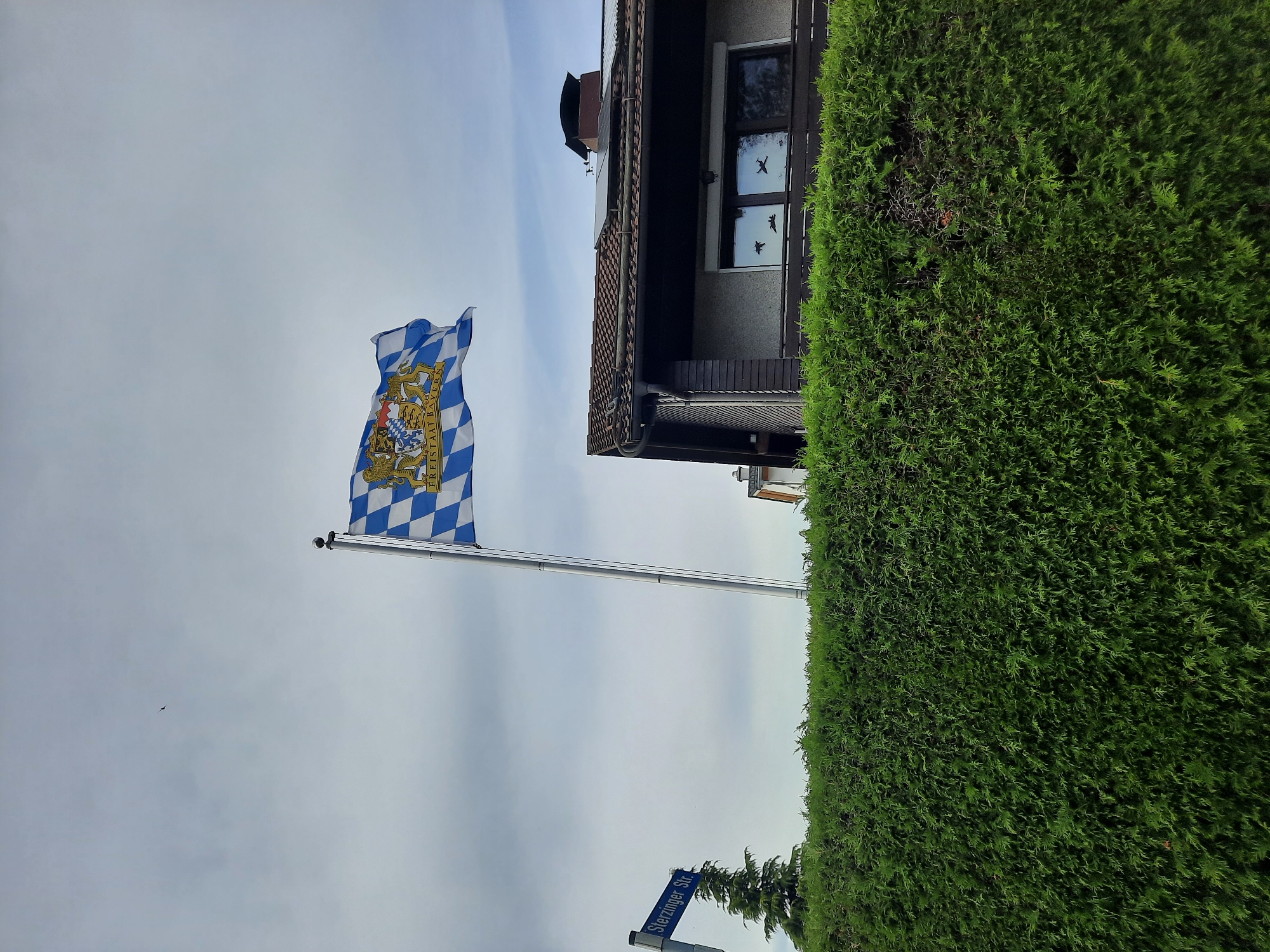 The image shows a flag with the coat of arms of Bavaria flying from a flagpole. The flag has a blue and white checkered pattern and the inscription "FREISTAAT BAYERN". A street sign that reads "STERZINGER STR." is visible to the left of the flagpole. Behind the flagpole and sign, a hedge is in the foreground. Beyond the hedge, a house with a brown tiled roof, solar panels, and a chimney is visible. The windows of the house have reflections of what appear to be fighter jets. The sky is overcast. The scene is located in Rosenheim, Germany, as indicated by the street sign.