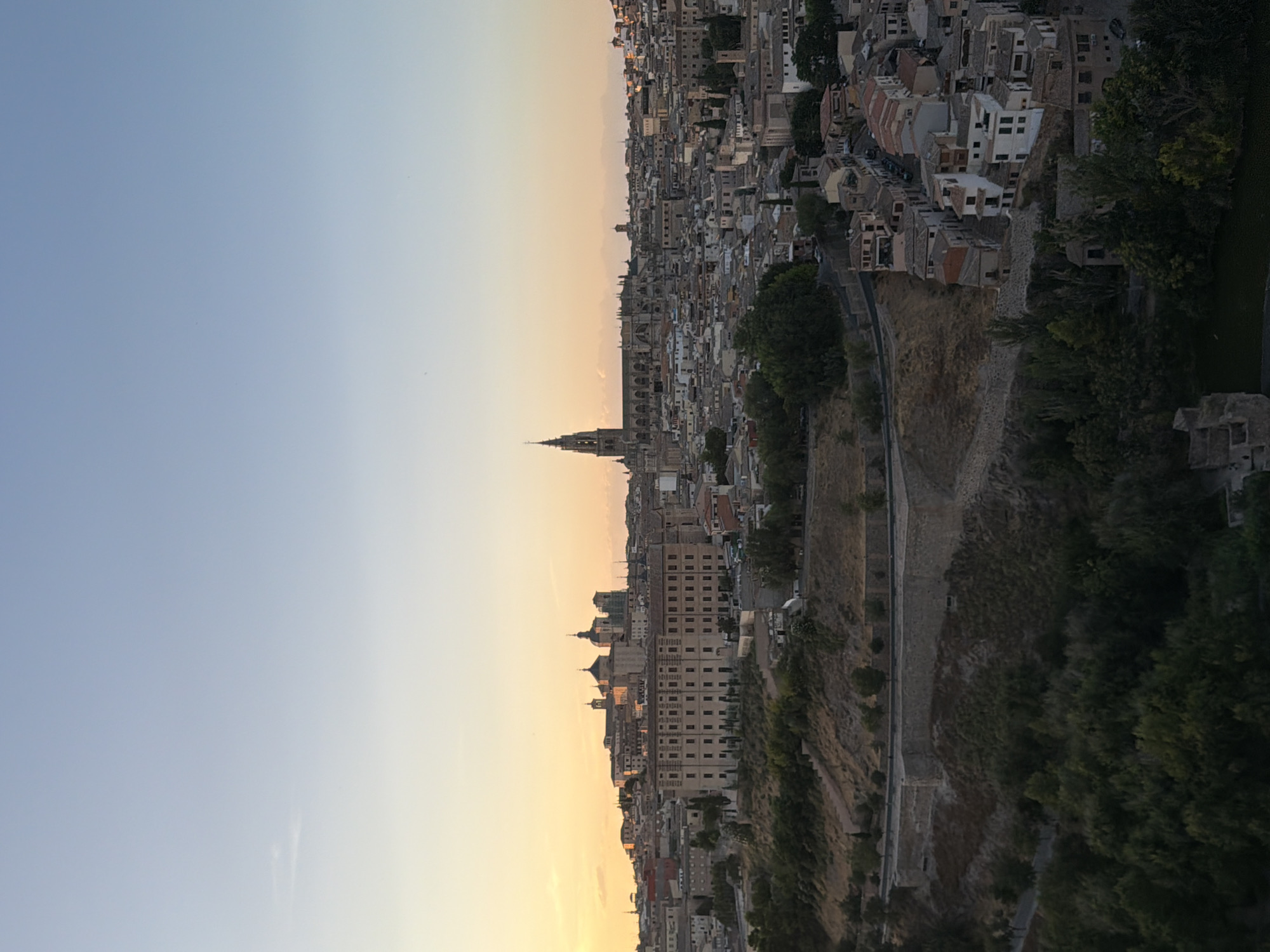 This is a high-angle, full shot of Toledo, Spain, taken at dusk. The sky transitions from a soft orange hue near the horizon to a pale blue overhead, with a few wisps of clouds. The city, built on a hillside, is densely packed with buildings of various sizes and architectural styles. Prominent structures include a large, long building with many windows in the foreground, and further back, a cathedral with a tall, spired bell tower. Other buildings feature red-tiled roofs and cream-colored walls. The landscape in the foreground is characterized by a steep, dry, grassy hillside with scattered trees and a stone retaining wall. In the very bottom of the image, a dark body of water, possibly the Tagus River, is visible, with the edges of some stone structures peeking out from its surface. There are no visible people or animals in the frame. The overall scene is one of a historic European city at the end of the day.