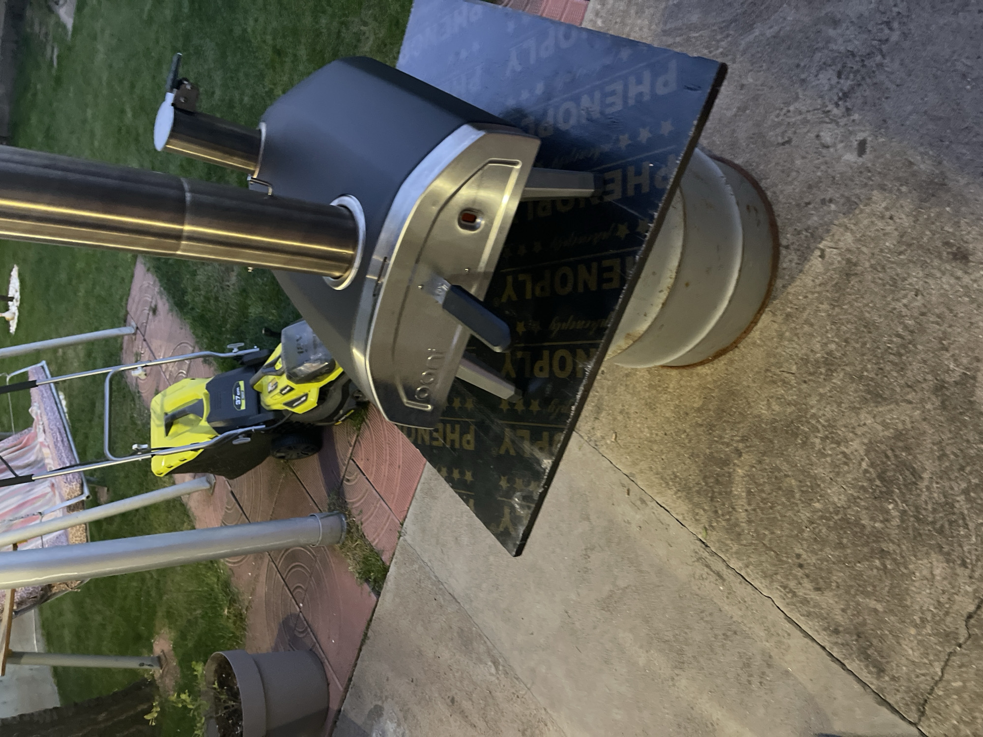 The image shows an outdoor scene, possibly a backyard or patio. A pizza oven, black and silver in color with a stainless steel chimney, sits on a wooden platform marked with the word "PHENOPLY" on top of a light grey, corrugated metal barrel. Adjacent to the oven is a yellow and black lawnmower. Behind the lawnmower is a beige and purple striped sun lounger. Nearby are a gray planter pot and parts of a metal sun lounger. The ground consists of grass, patterned pavers, and a concrete surface. There's no discernible evidence to place the scene in Sofia, Bulgaria.