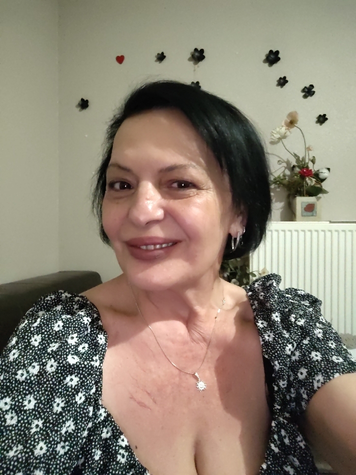 The image presents an indoor, chest-up portrait of a woman with dark, short hair, smiling directly at the camera. She wears a black garment with a white floral pattern and visible ruffled sleeves. Silver-colored hoop earrings are present on both ears, and a thin, silver-toned necklace with a sun-shaped pendant hangs around her neck. Superficial red linear marks are visible on the upper left side of her chest. The background features a light-colored wall adorned with a single red heart decal and multiple small, dark-colored flower-shaped decals. To the right, a white, ribbed radiator is partially visible, with a small vase containing artificial red and off-white flowers situated above it. A section of a dark brown or black sofa or chair back is discernible on the left side of the frame. The image contains no visual elements to confirm the specific location in the city of Ostwald, France.