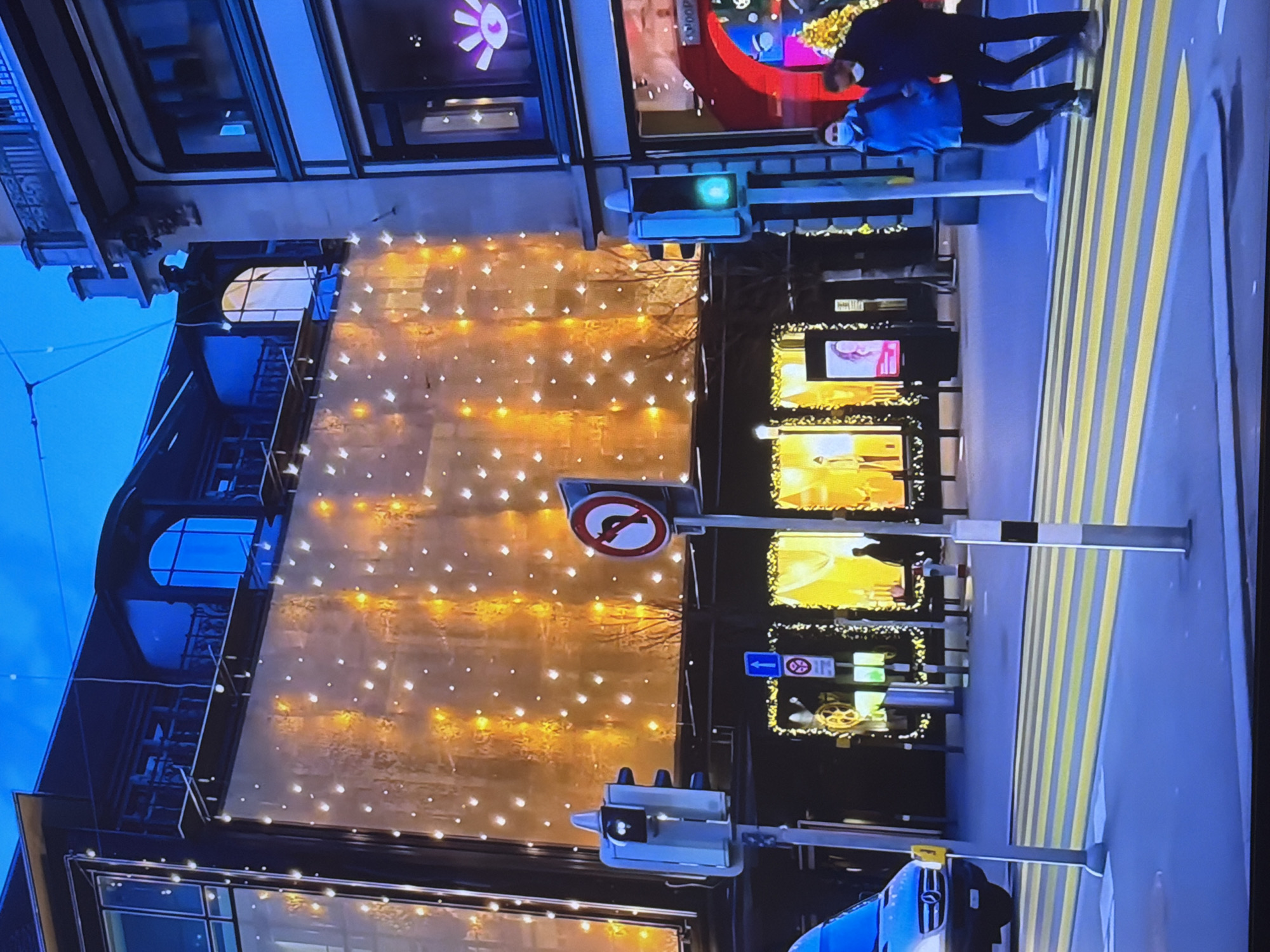 The image depicts a street scene at dusk or night in a city, likely in Europe given the architectural style of the buildings and the presence of traffic signals and signs. A large building with a facade covered in illuminated lights, possibly a department store or hotel, dominates the background. The lights are arranged in a scattered pattern, creating a festive or decorative effect. In the foreground, a pedestrian crossing with yellow and black stripes is visible. Two people, wearing masks, are walking across the street. Traffic lights are present, with one showing a green light. A van is partially visible on the left side of the frame. The sky is a deep blue, indicating twilight or nighttime. There is no indication of the city of Bac Nha Trang Ward, Vietnam in the image; the scene appears to be in a different geographical location.