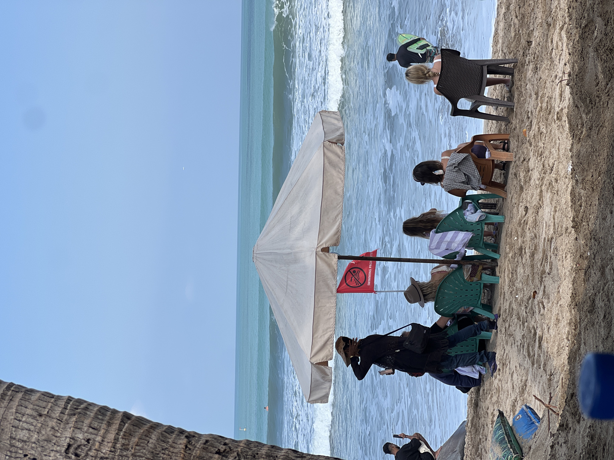 The image depicts a beach scene in Kuta, Indonesia. In the foreground, people are seated on chairs on a sandy beach, with a large umbrella providing shade. The ocean is visible in the background with moderate waves. A red flag with a prohibition symbol indicating "SWIMMING PROHIBITED" is planted in the sand. A person carrying a surfboard is walking into the water. A palm tree trunk is on the left side of the frame. The sky is mostly clear with some clouds.