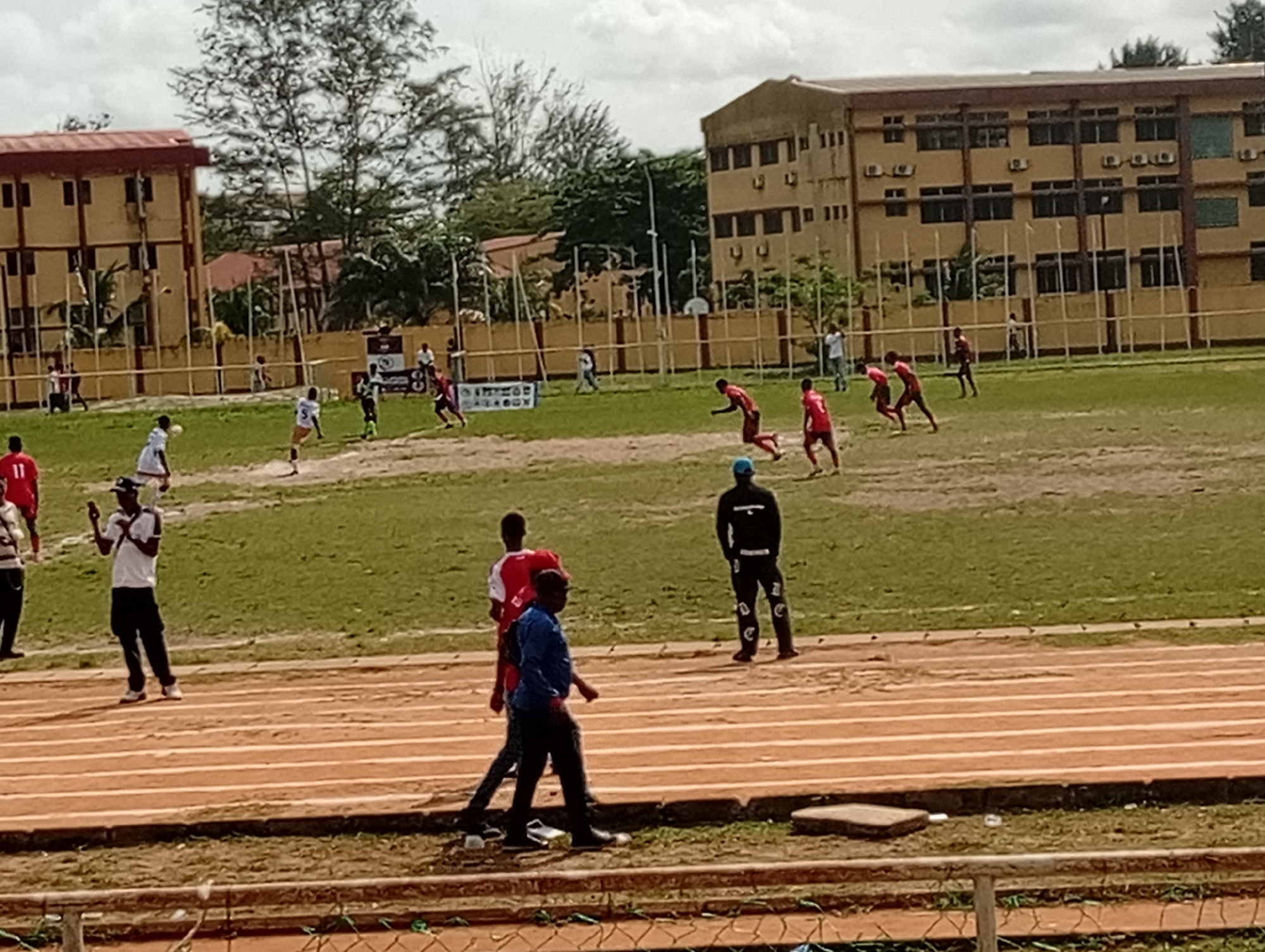A soccer game is in progress on a field in Ijanikin, Nigeria. Several players in red jerseys are running on the grass field, while others in white are defending. A few spectators are standing on the side of the field near a track marked with white lines. Buildings are visible in the background. The sky is overcast.