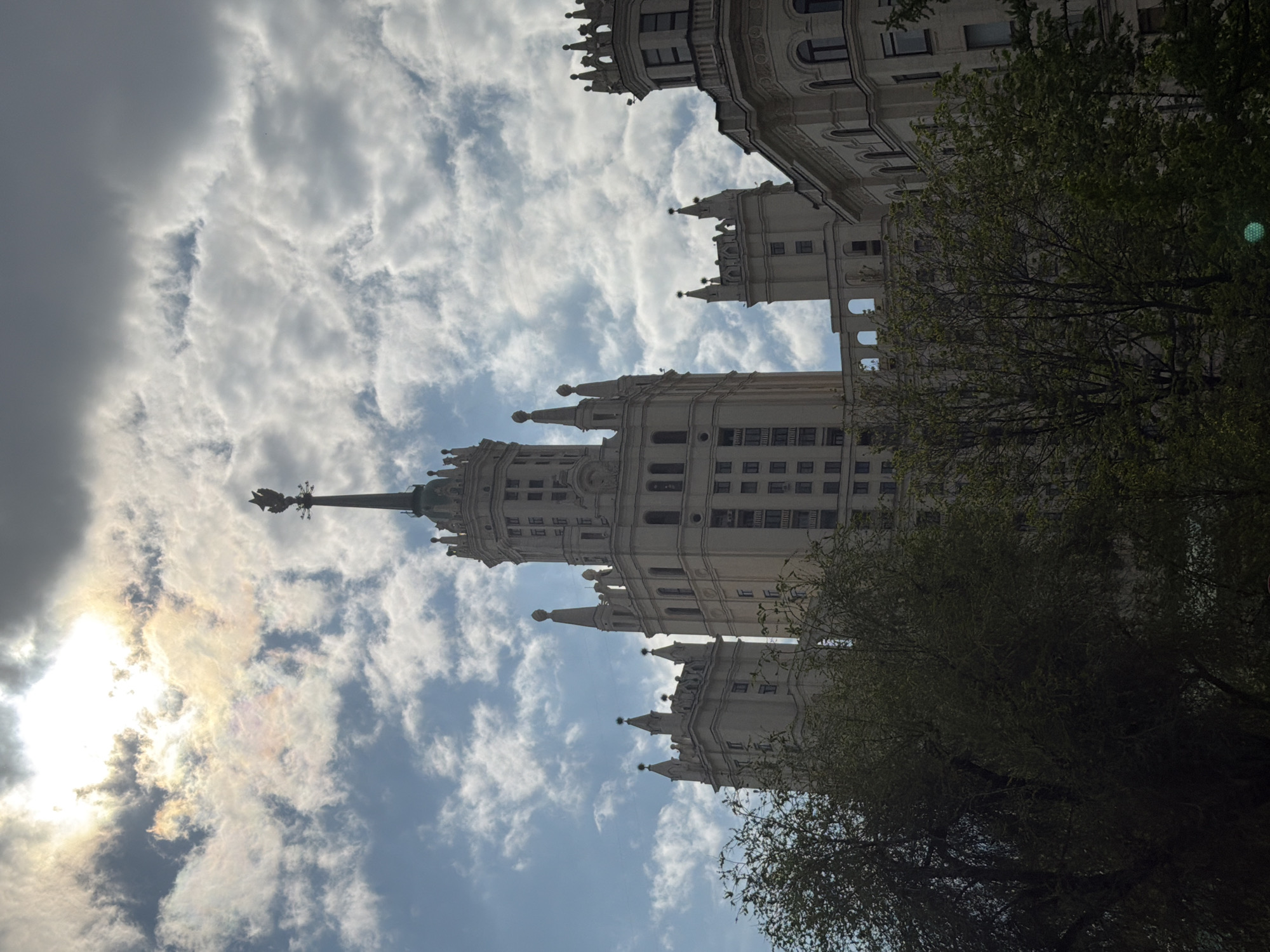 A low-angle shot depicts a tall, ornate building, partially obscured by trees on the right. The architecture features a slender spire and decorative detailing. The building is one of the Seven Sisters in Moscow, Russia. The sky is visible behind the building, with a mix of blue and cloudy weather. The sun is positioned to the left, obscured by a dark cloud.