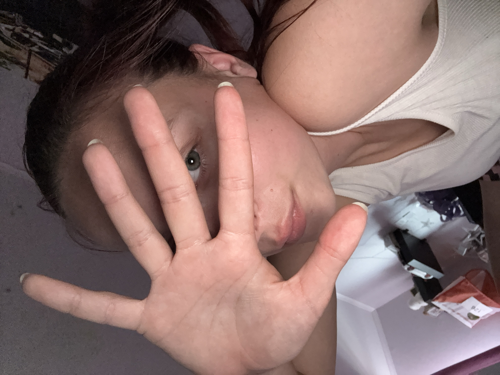 The image shows a close-up of a person's face and hand, obscuring most of their features. The person has dark hair and is wearing a white tank top. Their left hand is held up in front of their face, with fingers spread. The background appears to be a room with some objects in boxes, suggesting a domestic setting. The lighting is dim, indicating it might be nighttime or a dimly lit room. The image is taken from a low angle. No text is visible in the image. The scene implies the person is posing for a photo or is in the process of taking one.