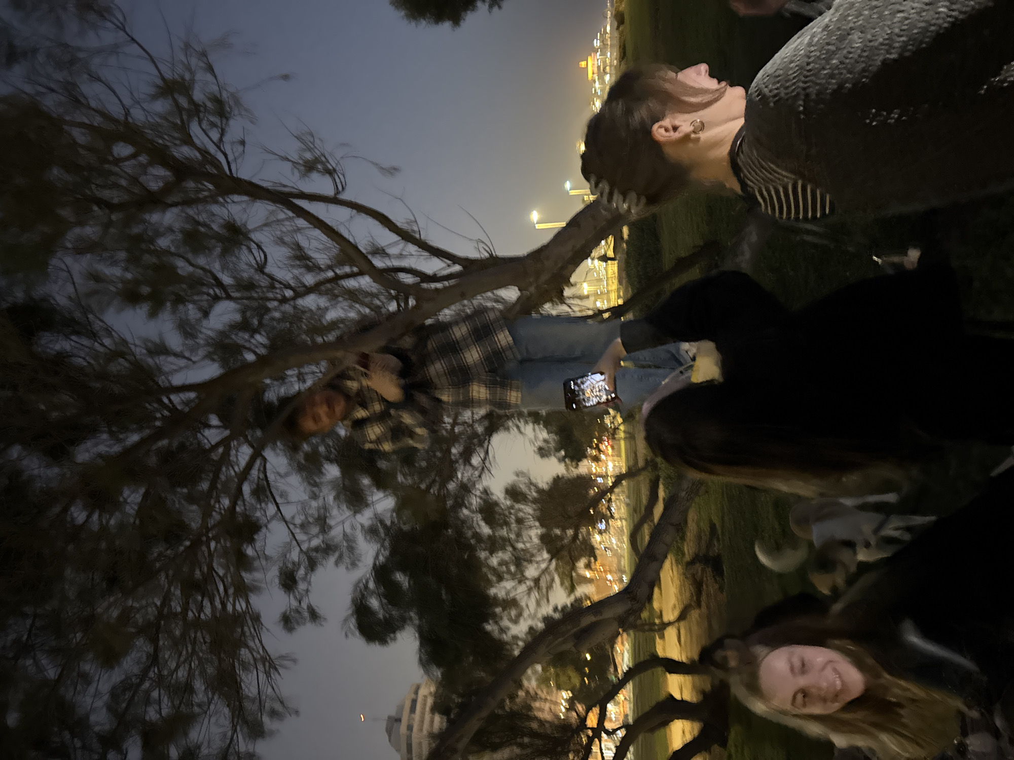The photo shows a group of people outdoors in Tel Aviv, Israel at night. A woman with long hair and hoop earrings is in the upper right of the image, looking towards the left. Next to her and slightly below is another woman with light-colored hair smiling. To the left, a person is holding up a cell phone and taking a photo. The background shows city lights and buildings, and there is a tree with many branches obscuring parts of the scene.