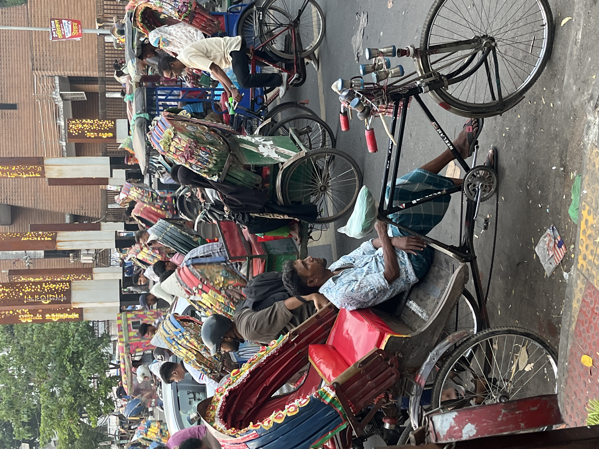 The image depicts a street scene in Dhaka, Bangladesh, dominated by numerous cycle rickshaws and their drivers. Many rickshaws are adorned with colorful decorations. Drivers are either resting in or near their rickshaws, waiting for fares. The environment includes urban infrastructure, such as buildings and streetlights with Bengali signage. The overall scene suggests a typical busy day in Dhaka, showcasing a key mode of transportation.