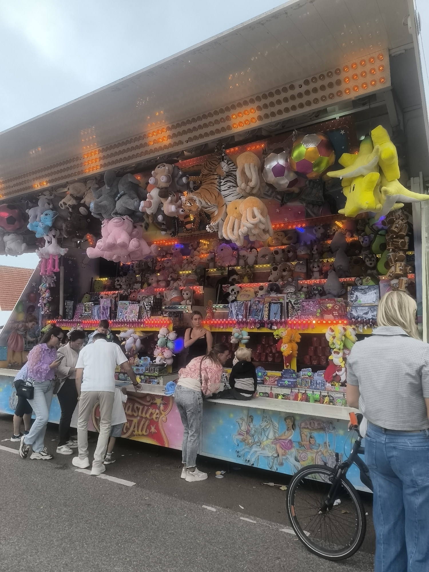 A fairground game stall filled with plush toys is displayed under an overcast sky in Onderdijk, Netherlands. The stall features rows of stuffed animals, including tigers, elephants, octopuses, and various cartoon characters, hanging from the ceiling and arranged on shelves. Brightly colored lights line the front of the stall, illuminating the prizes. Several people are gathered at the stall, some appearing to play the game while others are observing. A bicycle is parked in the foreground, leaning against the stall. The ground in front of the stall is paved with asphalt, and a white line marks a boundary. The overall scene suggests a lively atmosphere typical of a fair or amusement park.