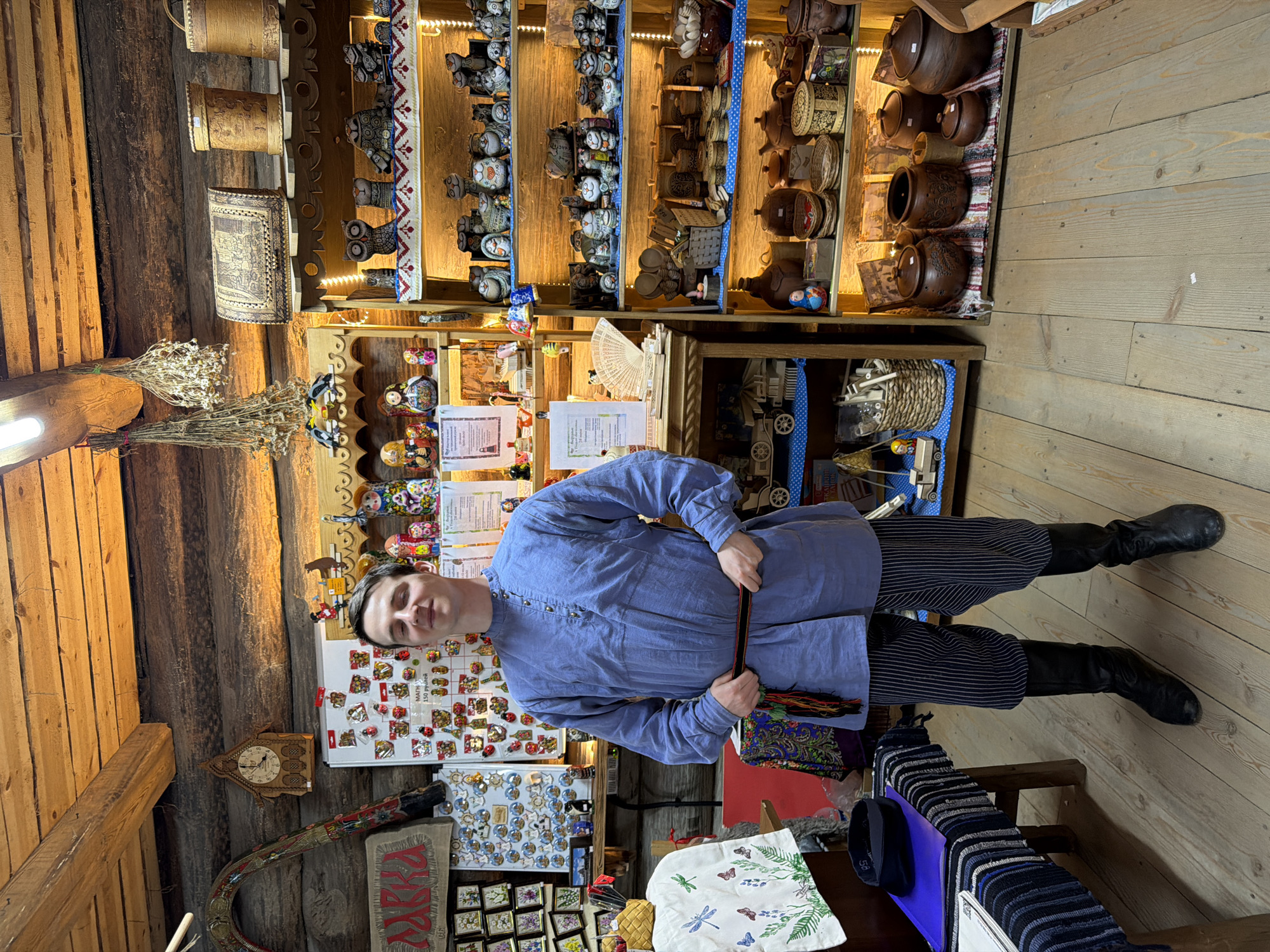 A man stands in a wooden room filled with traditional Russian crafts. He is wearing a light blue, long-sleeved shirt with a Mandarin collar and a dark striped sash around his waist. He also wears dark striped pants and black leather boots. The room is decorated with shelves displaying a variety of items, including nesting dolls, ceramic figures, wooden boxes, pottery, and woven baskets. A clock hangs on the wall behind the man, and dried herbs are suspended from the ceiling. The overall impression is that of a shop or museum selling folk art in the city of Imeni Sverdlova, Russia.