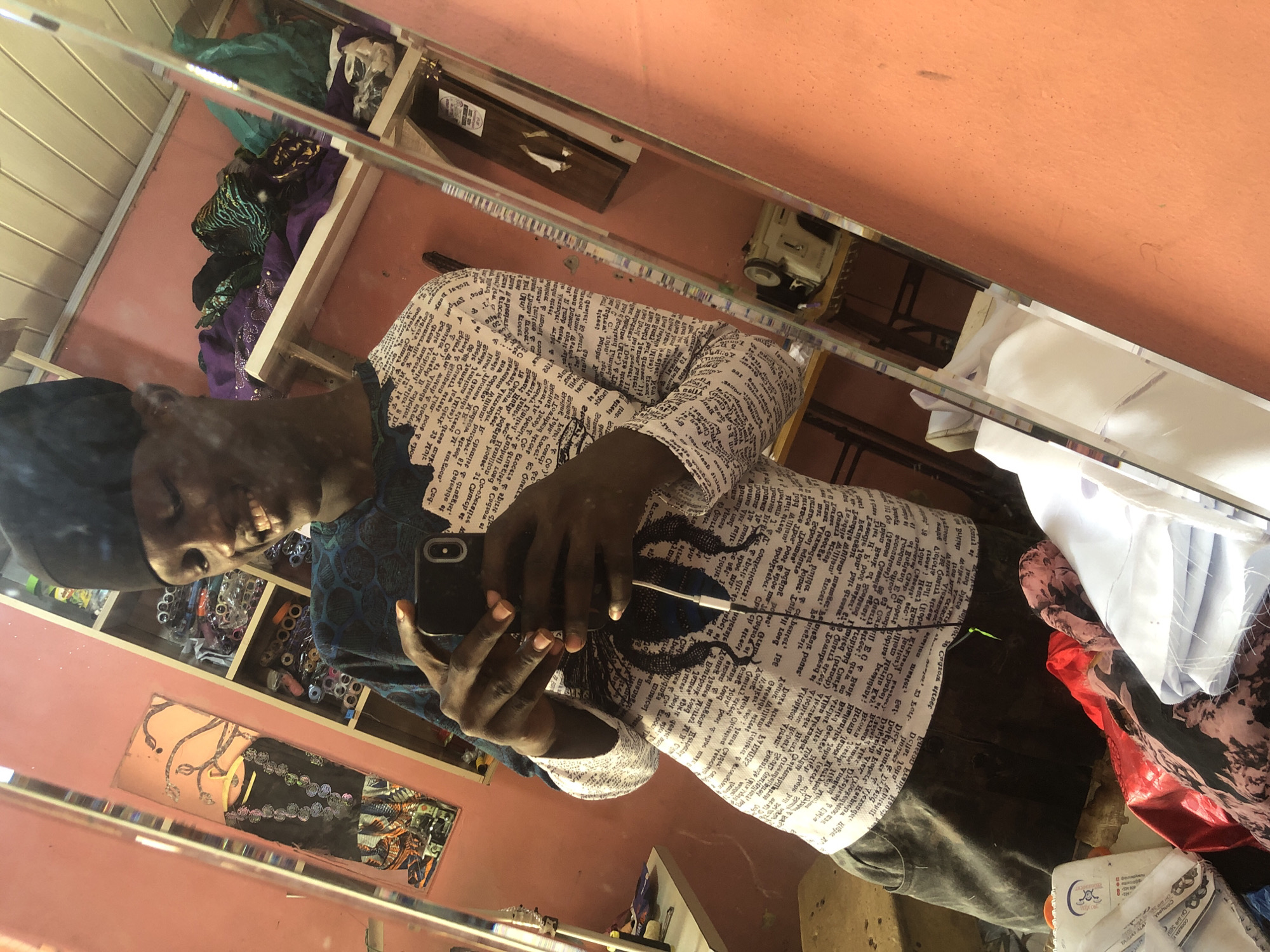 A young Black man wearing a black skullcap and a long-sleeved shirt with a black and white text print is taking a selfie in a mirror. He is smiling and holding a smartphone in his right hand, pointed towards the mirror. The background appears to be a tailor's shop, likely in Ado, Nigeria. Shelves filled with fabrics, sewing supplies, and a sewing machine are visible in the reflection. The walls are painted a peachy-pink color. A table with some fabric remnants is also present. The overall impression is a casual moment captured in a working environment.