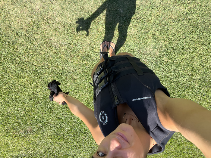 A young adult female is depicted in a high-angle selfie, standing outdoors on a manicured green grass surface in Coruche, Portugal. She wears a black NEILPRYDE impact vest or life jacket, featuring a central zipper and multiple horizontal buckle straps. A layered gold-colored necklace is visible around her neck. In her right hand, she holds a crumpled black fabric object. Her feet are clad in black flip-flops, and the sunny conditions cast distinct shadows of her lower body and arm onto the grass. She looks directly at the camera with a slight smile.