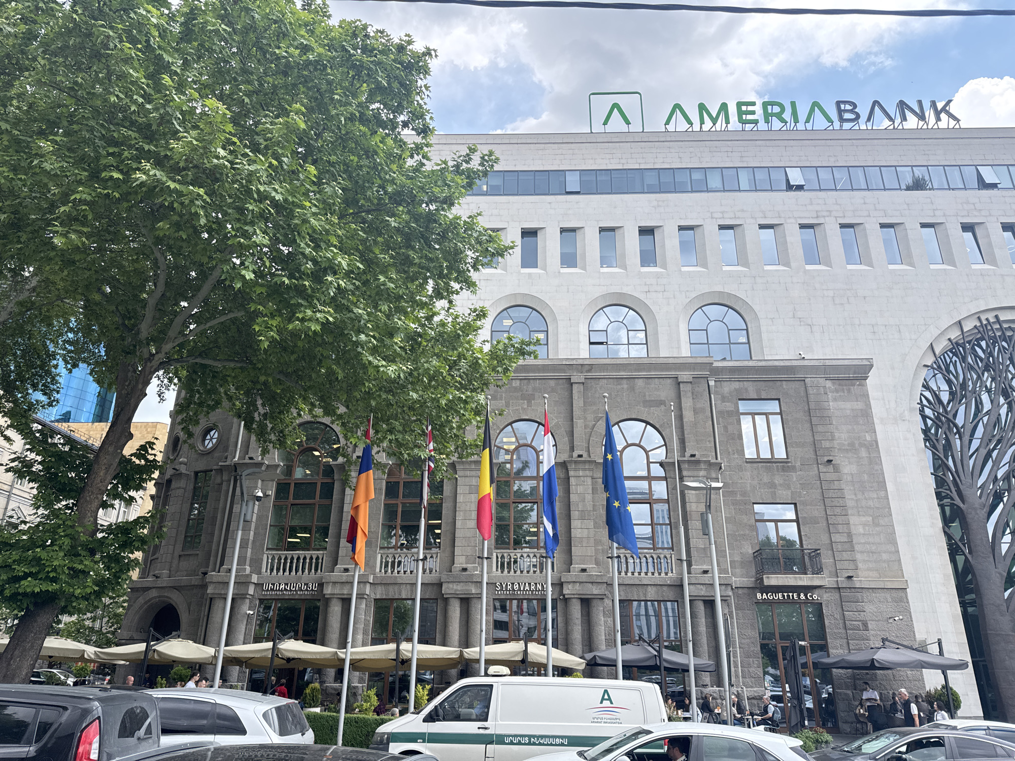 The image shows a street view in Yerevan, Armenia, featuring the AmeriaBank building. The building has a layered facade, with an older, stone-clad lower section and a modern upper section with a white facade and the AmeriaBank logo in green on top. Several national flags, including the flags of Armenia, the United Kingdom, Belgium, Netherlands and the European Union, are displayed in front of the building. Below the flags, there are several businesses; "Syrovarny Eatery-Cheese Factory" and "Baguette & Co." are visible. A white van with green stripes and an "A" logo is parked in the street, along with other cars. People are seated at tables outside the "Baguette & Co." shop. A large tree on the left side of the image partially obscures the view of the building. The sky is cloudy.