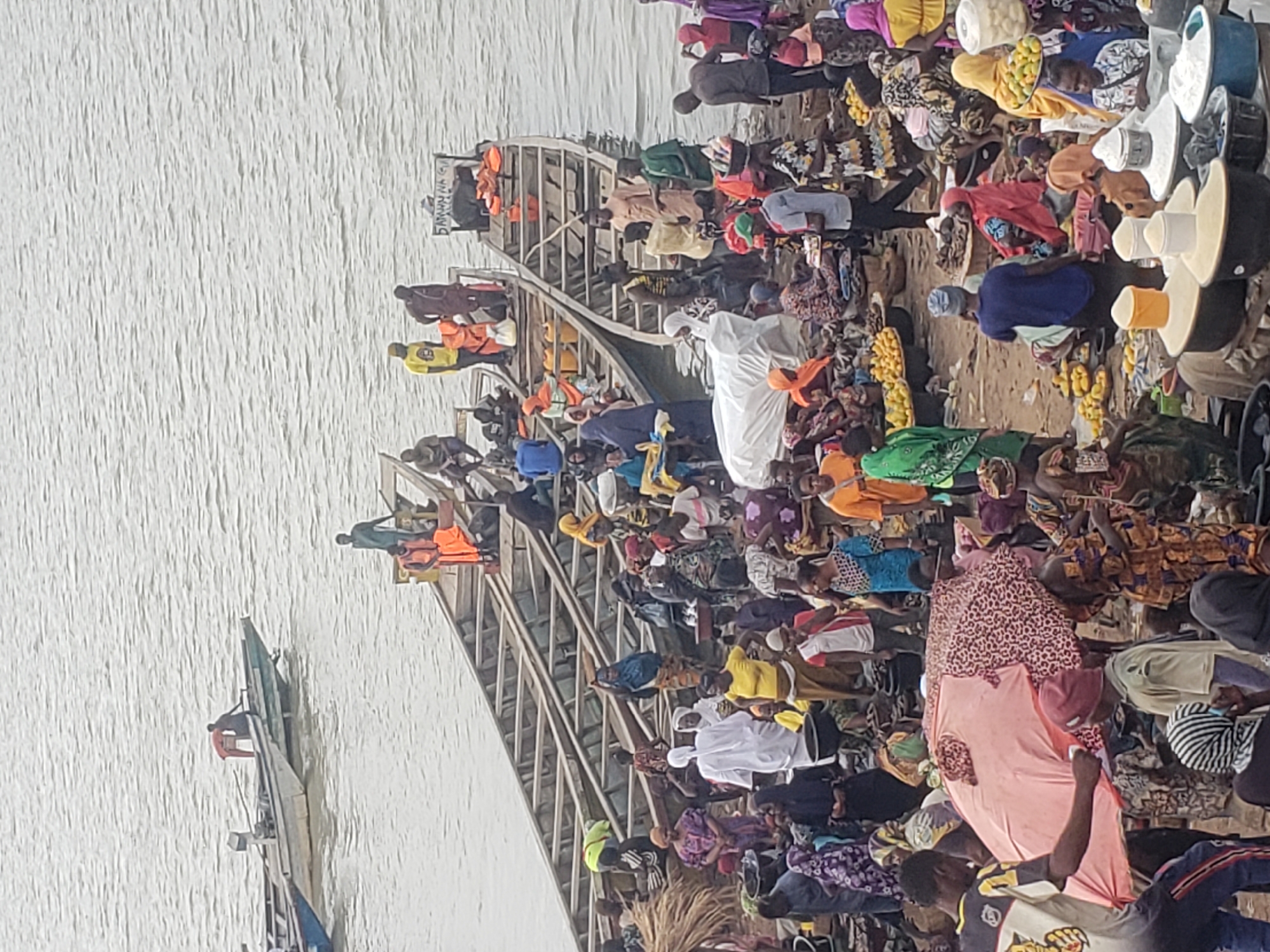The image captures a bustling daytime scene at a riverbank in Lokoja, Nigeria, packed with a large crowd of people, several boats, and various goods, suggesting a vibrant market and transportation hub.
The foreground and midground are densely populated with individuals of all ages, dressed in diverse, colorful clothing common in West Africa. Many people are actively engaged in moving around, standing, or sitting. Notably, several individuals, particularly those near or on the boats, are wearing bright orange and yellow life vests, indicating preparation for river travel.
Multiple long, wooden boats, resembling ferries or large canoes, are docked or positioned near the bank. The largest boat in the center features multiple seating levels or decks, appearing to be actively loaded or unloaded with passengers and goods. Another smaller boat is visible further out on the water to the left.
The sandy bank is filled with various items for trade. Large baskets and white containers are prominently displayed, some overflowing with yellow fruits, likely mangoes or plantains, and other goods, signifying a marketplace. People are seen interacting around these goods, suggesting commercial transactions.
The overall activity portrays a busy point of embarkation and commerce, highlighting the importance of river transport and local markets. The natural, bright lighting indicates it is daytime. On the side of one of the boats, partial text "SABAHAN NA G..." is visible. The scene conveys a strong sense of community activity and economic exchange along the water.