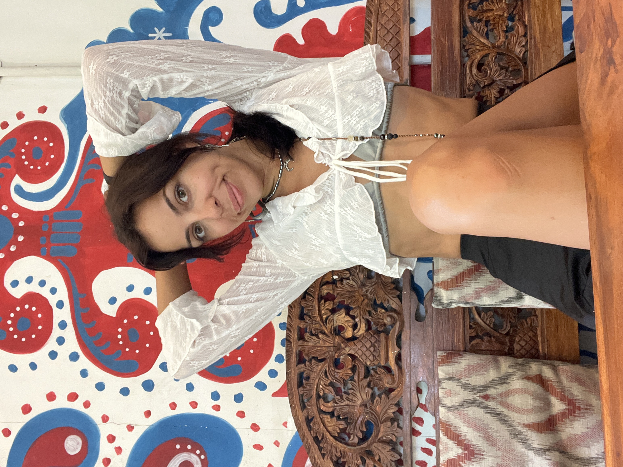 A woman with dark hair is lying on an ornate wooden chair. Her arms are behind her head, and she is looking at the camera with her tongue slightly out. She is wearing a white, sheer, ruffled blouse and denim shorts. She also has a necklace with a crescent moon charm.
Behind her is a wall painted with red and blue patterns on a white background. The chair she is lying on has intricately carved wooden panels, and there are patterned cushions underneath her. The environment suggests she may be in Kuta Selatan, Indonesia.