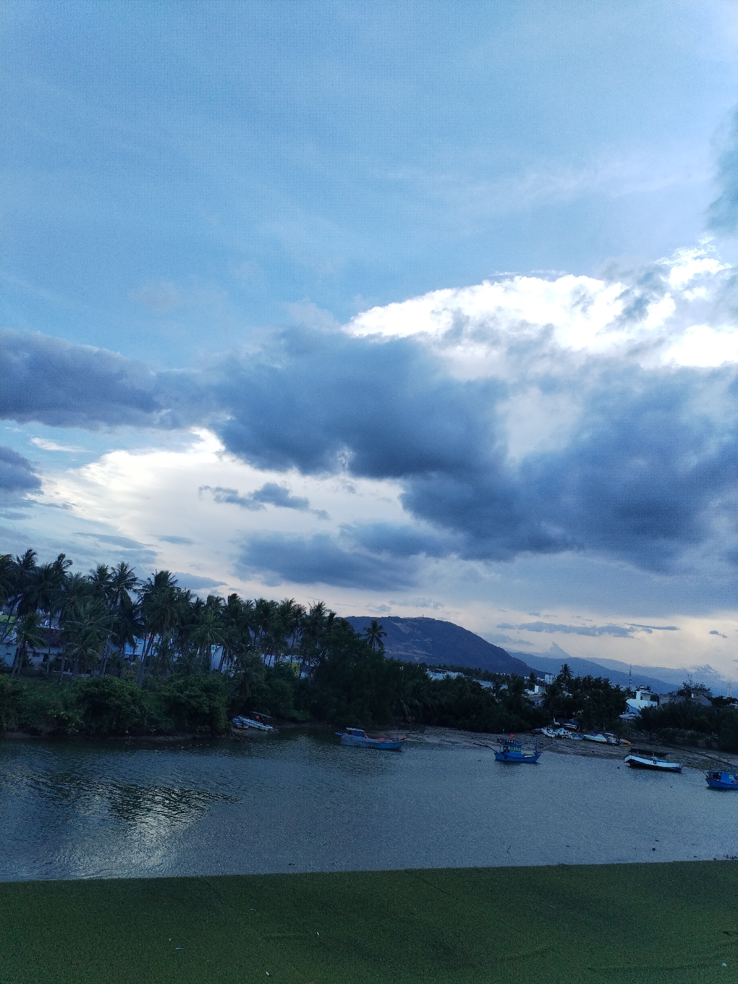 The image depicts a waterfront scene in Bac Nha Trang Ward, Vietnam, under a cloudy sky. In the foreground is a body of water, possibly a river or bay, with calm surface reflections. Several small fishing boats are docked along the shoreline. Behind the water is a line of trees, including palm trees, which suggest a tropical environment. In the distance, a mountain range is visible. The overall atmosphere suggests a coastal town setting with a blend of natural and urban elements.