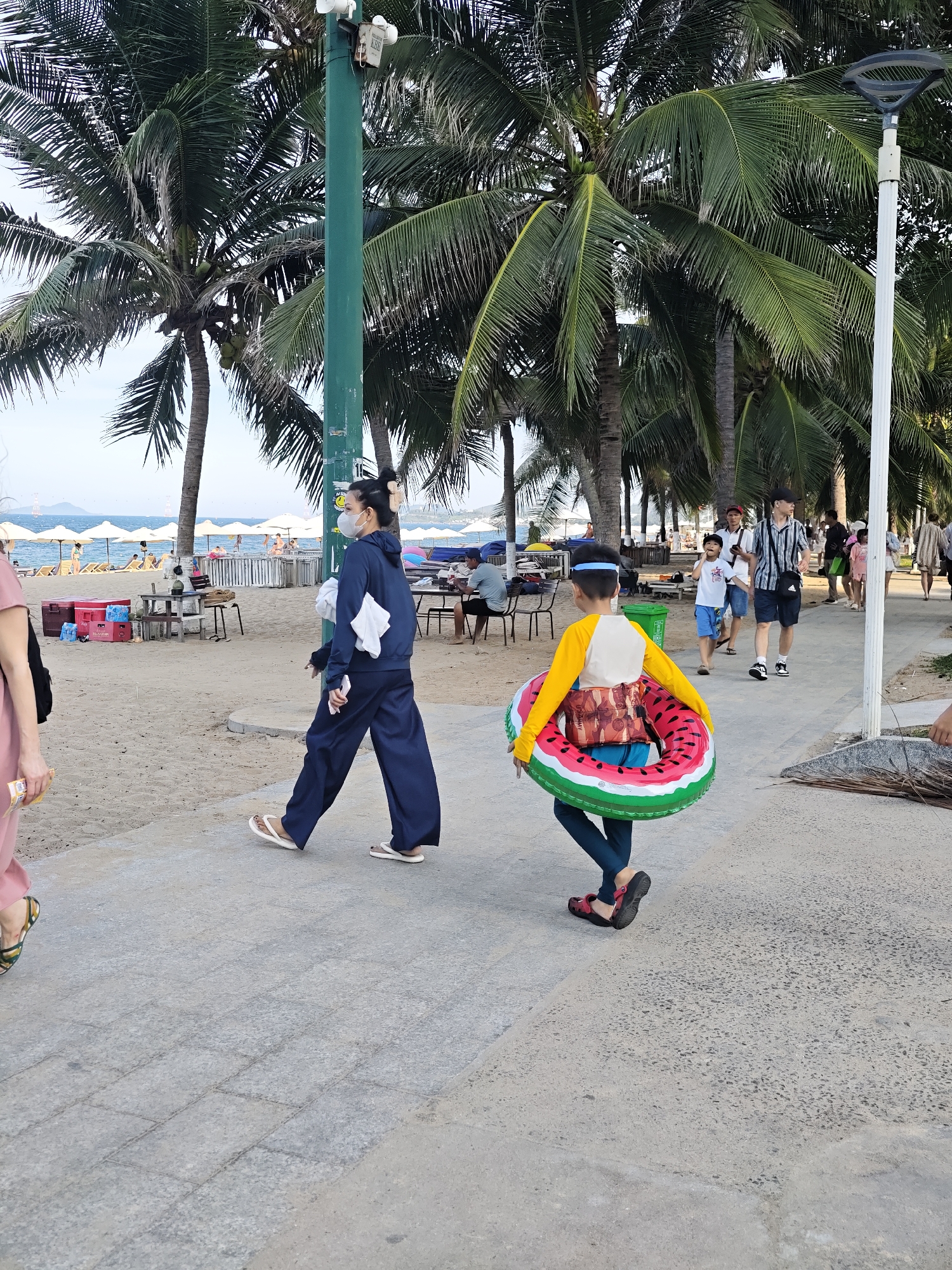 A full-shot, eye-level, outdoor, daytime, street-level photograph captures a scene on a paved walkway bordering a sandy beach in Nha Trang Ward, Vietnam. Palm trees line the background, with the ocean and beach umbrella-covered loungers visible beyond. In the foreground, a woman wearing a blue hooded jacket, dark blue pants, and white flip-flops walks on the paved path, carrying a white cloth in her right hand and a smartphone in her left. To her right, a child wearing a yellow long-sleeved shirt, blue shorts, and red sandals walks ahead, carrying a large, circular watermelon-patterned inflatable ring. Other pedestrians and beachgoers are visible in the middle ground and background, enjoying the seaside environment. The lighting suggests it is late afternoon or early evening.