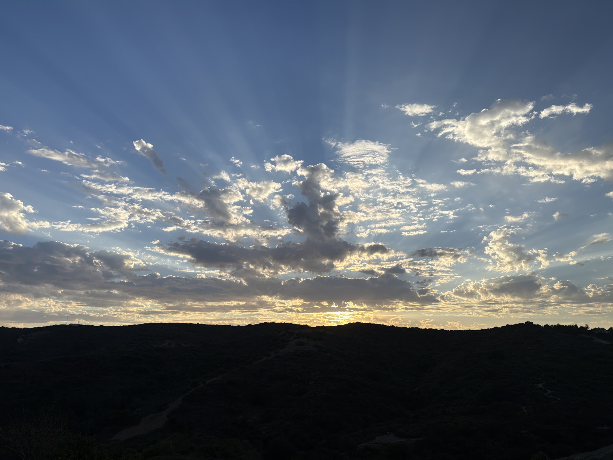 A wide, horizontal, eye-level shot captures a sunset over a silhouetted, rolling landscape in Aliso Viejo, California. The sky is a gradient of deep blue at the top, transitioning to lighter blue and then a pale yellow-orange near the horizon, where the sun is setting. Rays of light fan out from the sun, piercing through scattered, fluffy clouds that are illuminated by the setting sun. The clouds are white and gold, with some darker gray undersides. The foreground is dominated by a dark, silhouetted hill or mountain range, obscuring any details of the land below. The undulating silhouette suggests a natural, rugged terrain. No people or artificial structures are clearly visible, lending the scene a sense of natural beauty and tranquility.