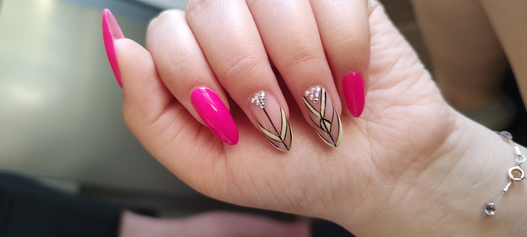 A close-up shows a person's left hand with painted fingernails. The nails are painted in bright pink and beige colors, with black line designs and small rhinestones on the beige nails. The hand is resting on a surface, and a bracelet with small clear beads is visible on the wrist. The location of the image is not specified.