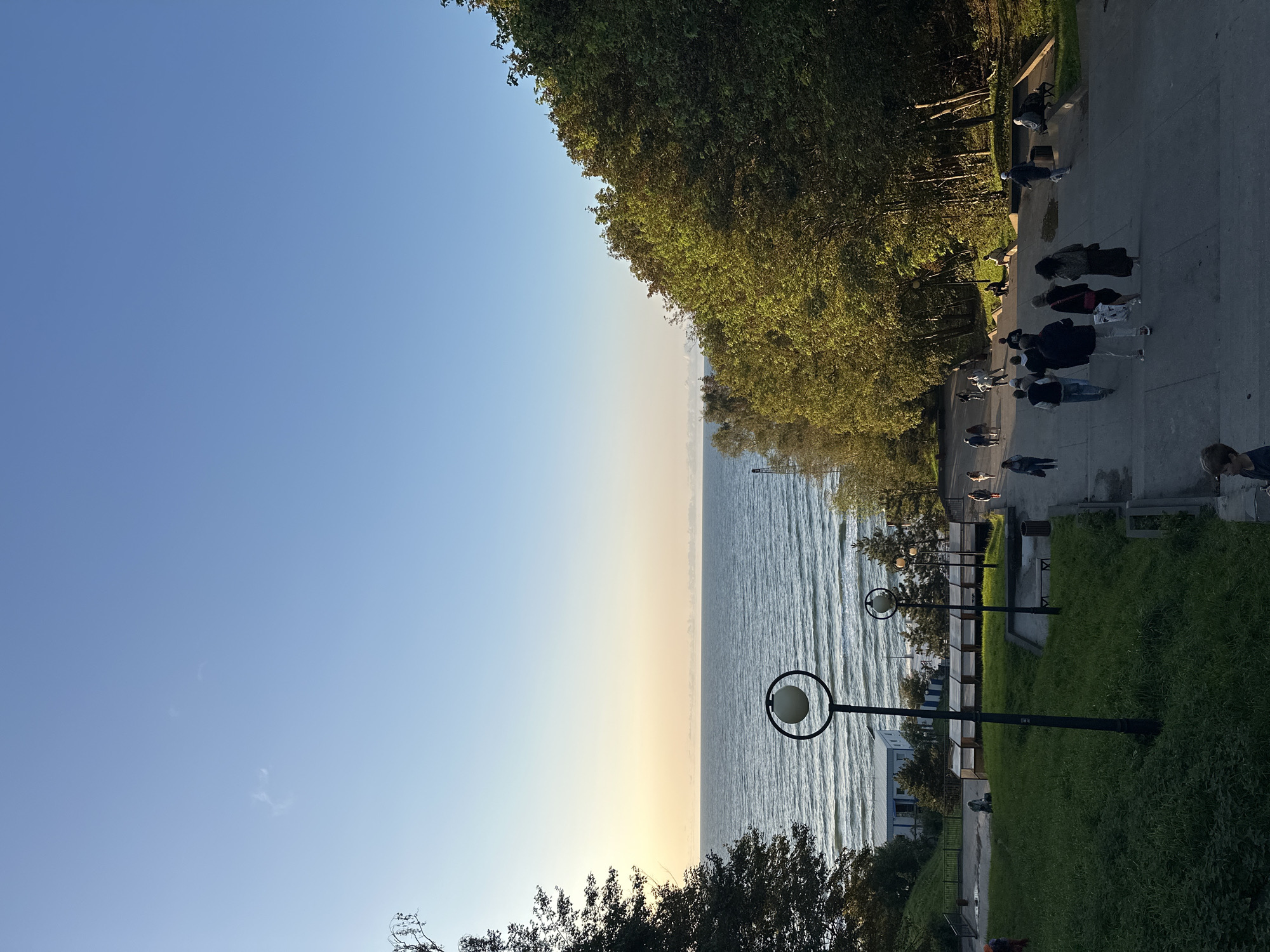 The image depicts a wide outdoor staircase leading down towards a body of water, likely the Baltic Sea, in Svetlogorsk, Russia. The sky is clear and blue, with a hint of pale yellow near the horizon, suggesting either sunrise or sunset. Lush green trees flank the right side of the staircase, with some trees also visible on the left. Several people are walking up and down the stairs, dressed in casual attire. A few streetlights are positioned along the path. In the distance, near the water's edge, there appears to be a small building and some beach structures. The overall scene conveys a peaceful, outdoor setting.