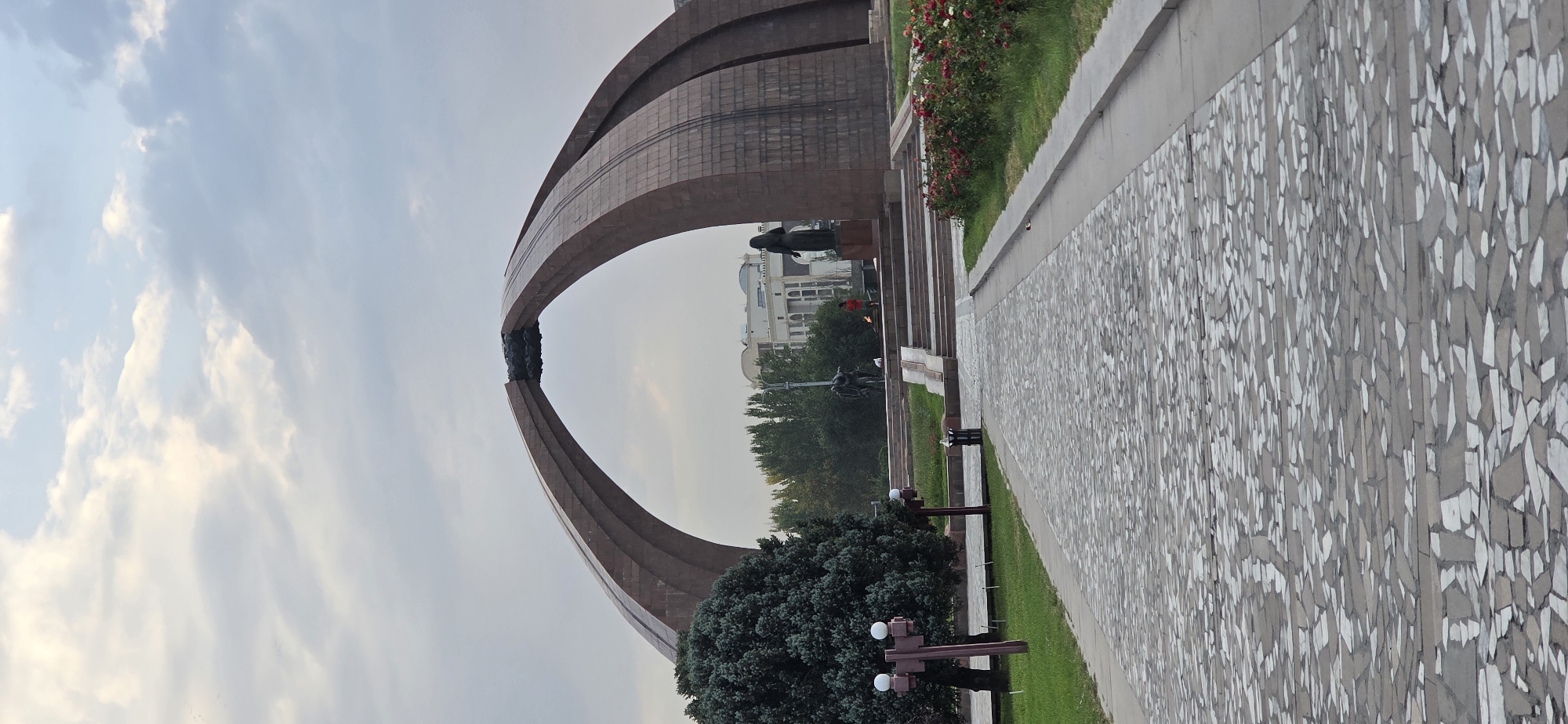 The image shows a monument in Bishkek, Kyrgyzstan, with a wide, mosaic-paved pathway leading towards it. The monument is an arched structure made of large, reddish-brown stone blocks, with a dark, ornate element at the apex of the arch. To the left of the pathway is a large, dark green evergreen tree, and two lampposts with spherical lights. In the background, behind the monument, are trees and modern buildings. There are also statues and a flame of remembrance visible on a raised platform leading up to the monument. A few people are present, including one wearing a red shirt standing near the monument, and two figures in bronze statues. The sky is overcast with soft, diffused light.