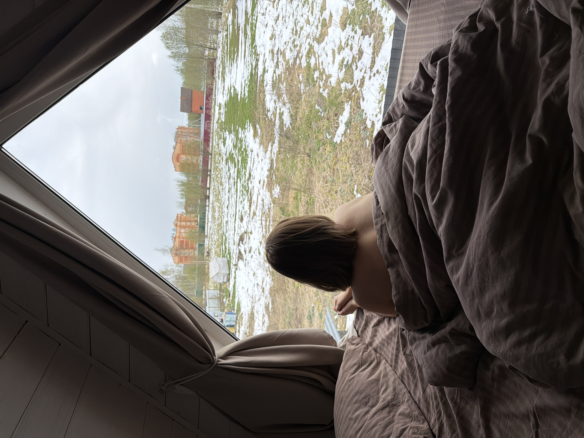 The image captures an indoor perspective, looking out a large window, with the scene rotated counter-clockwise. A person, with their back and the back of their head visible, lies on a bed under a dark grey or brown duvet, seemingly leaning forward to look out the window. The bed is covered in crumpled, dark fabric.
Through the white-framed window, an outdoor scene unfolds under an overcast, grey sky. The ground is a patchwork of melting snow, dry brown grass, and emerging green patches, indicative of late winter or early spring conditions. Sparse, bare trees are scattered across the landscape. In the distance, several buildings are visible, including a small reddish-brown house with a dark roof, and larger, multi-story residential buildings with orange-toned facades.
Light beige curtains are tied back to the sides of the window, and a white paneled wall is visible below the window frame. The overall atmosphere suggests a quiet, contemplative moment, possibly in the morning, observing the outdoor environment from a warm interior. There is no visible text in the image. The location context is Бунятино, Russia.