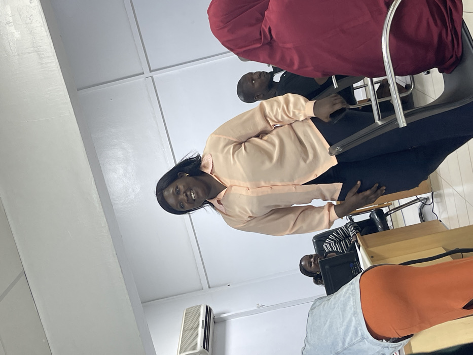 The scene depicts an indoor gathering of several individuals in a brightly lit room. The location is specified as the City of Zaria, Nigeria.
A dark-complexioned adult female, with dark hair, stands prominently in the mid-ground, slightly off-center to the right. She is wearing a light peach-colored long-sleeved button-up shirt with a ruffled collar and dark trousers. Her expression is a broad smile, and she is looking towards the left side of the frame. Her right hand rests on the back of a black office chair.
To her left, partially obscured by a light blue denim jacket and an orange skirt worn by a person in the immediate foreground, a dark-complexioned female is seated at a light brown wooden desk. This seated female, wearing a black and white striped shirt, is also smiling and looking towards the left, with a black computer monitor visible on the desk in front of her.
To the right of the central standing figure, a dark-complexioned male with short hair is partially visible, seated in a dark shirt, also looking towards the left. In the extreme foreground on the right, the back and right side of a person wearing a maroon head covering and garment are visible, seated on a black chair.
The room features plain white walls, a white ceiling with visible trim, and a light-colored tiled floor. A white wall-mounted air conditioning unit is present on the upper left wall. Several black office chairs with metal frames are visible throughout the space. The overall environment suggests an office, classroom, or meeting room setting.