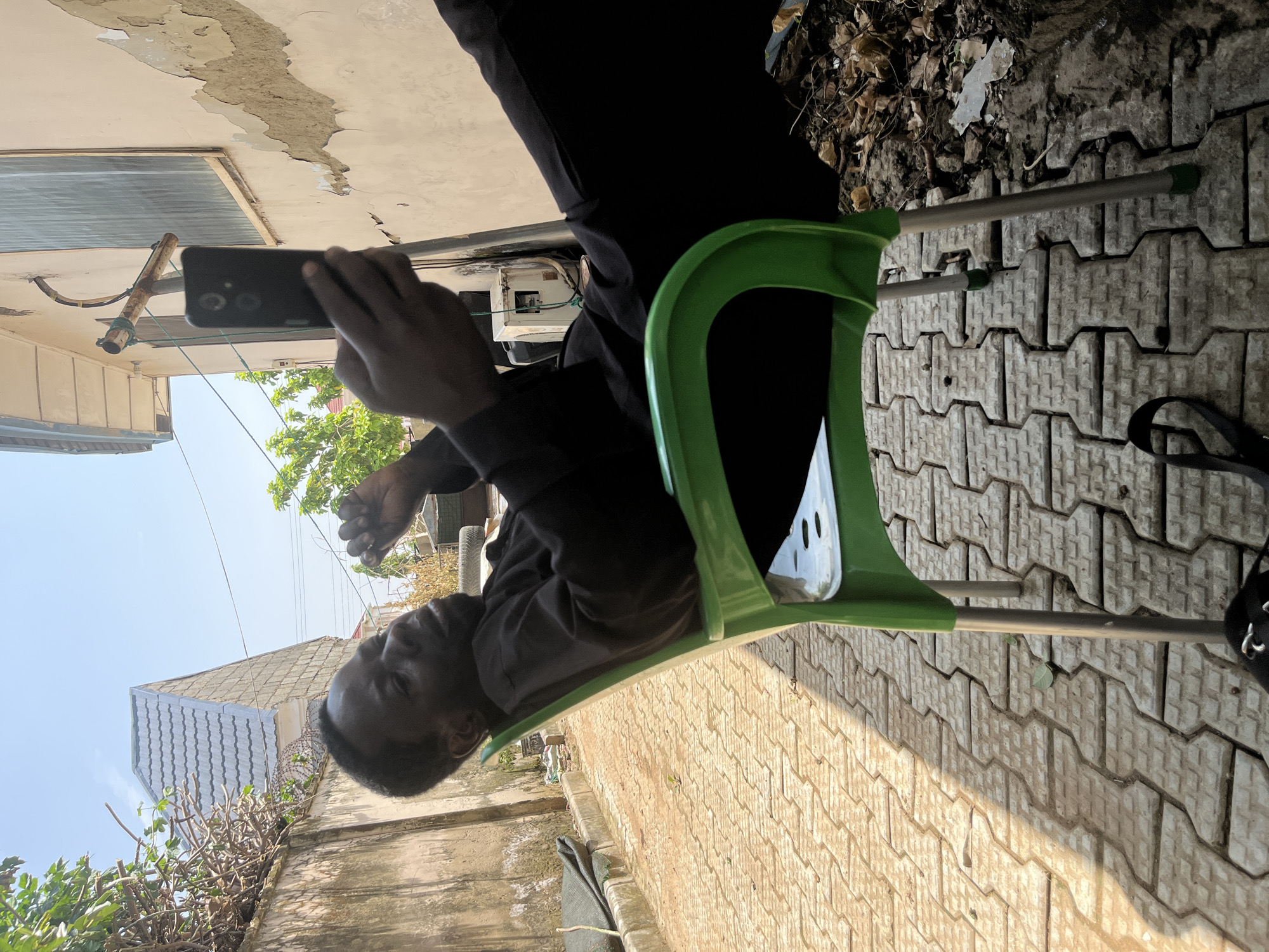 A man is sitting on a green plastic chair in what appears to be an outdoor area in Abuja, Nigeria. He is wearing black clothing and holding a smartphone in his right hand, seemingly looking at the screen. The chair has metal legs and is placed on a patterned paved surface. Behind him, a building with a light-colored, weathered facade is visible, along with some greenery and other objects on the ground that suggest an urban environment. The lighting indicates it is daytime, with shadows cast on the paved ground.