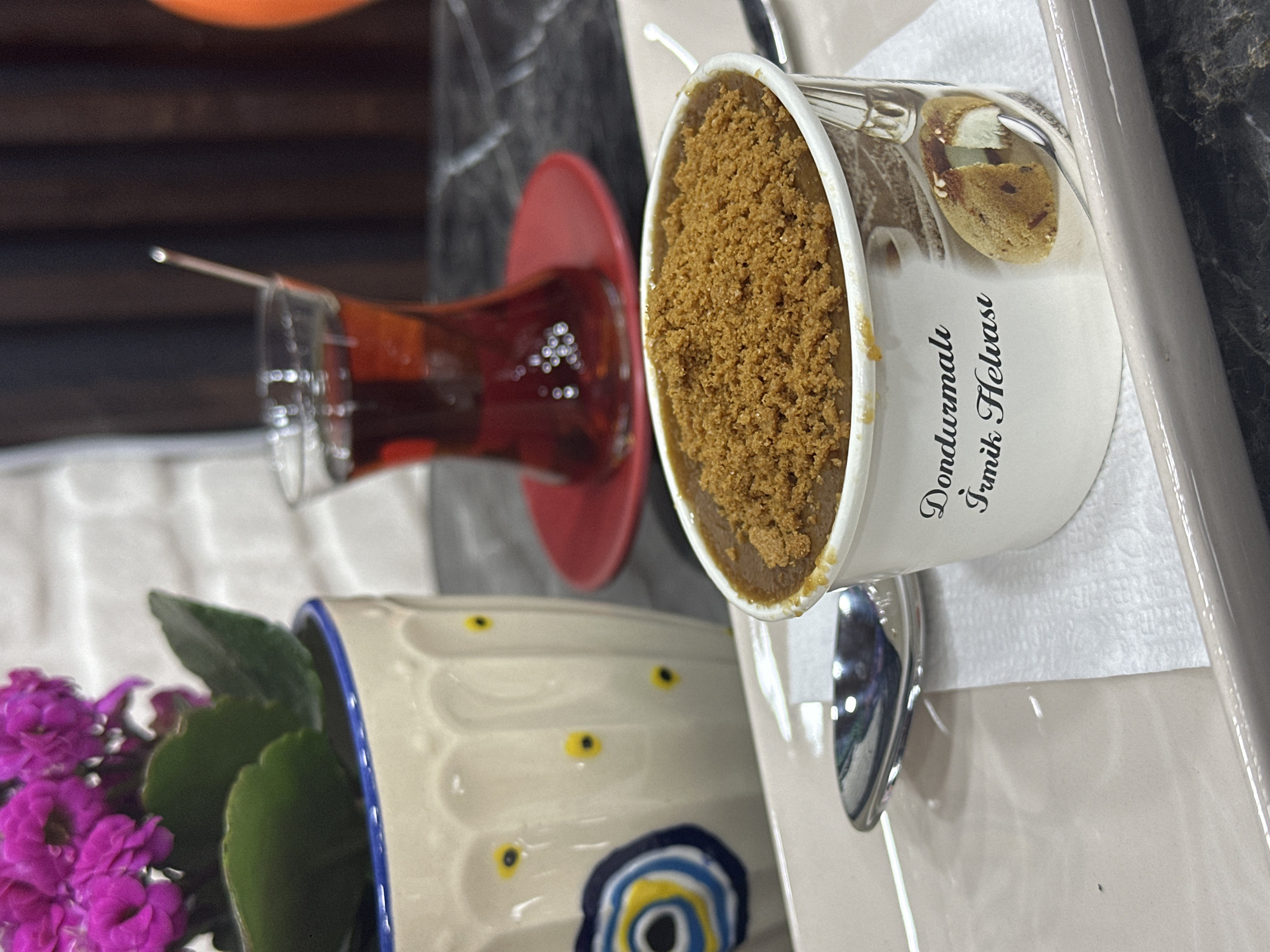 A table scene includes a glass of tea on a red saucer, a container of "Dondurmalık Irmik Helvası," a pot of purple flowers, and a spoon. The photo likely originates from Datça, Turkey, given the Turkish writing on the helva container.