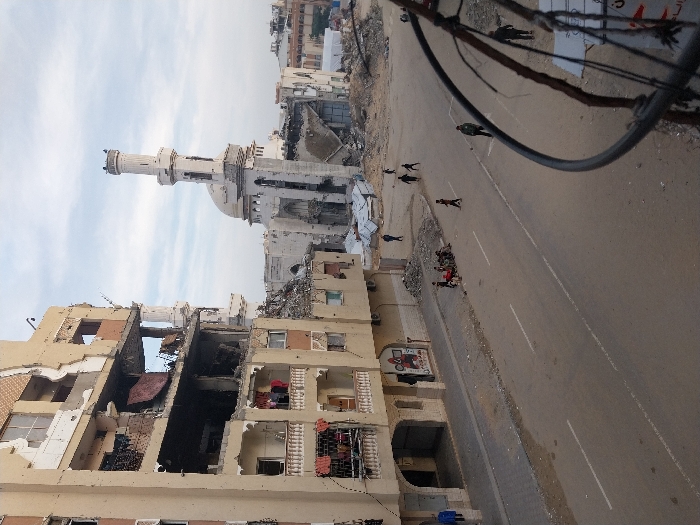 The image captures a wide street view in Khan Yunis, Palestinian Territory, depicting extensive urban destruction under an overcast sky. To the left, a multi-story building, appearing to be residential, shows significant structural damage with large sections of its beige facade missing, exposing multiple floors and interiors. Impact marks are visible across its remaining walls, and items of clothing (red, white, blue) are hanging from damaged balconies and exposed rooms.
In the mid-ground and right, the damaged remains of a large white mosque are prominent, featuring a tall, slender minaret that remains partially upright but visibly damaged, alongside a partially collapsed dome and surrounding structure. A substantial accumulation of rubble lies at the base of the mosque.
A paved asphalt road, featuring faded white lane markings, spans the mid-ground. The adjacent sidewalks and open areas are covered with dirt and scattered debris. Approximately 10-15 individuals, appearing to be a mix of children and adults, are present in the scene. A concentrated group of 6-8 individuals, mostly children, are seated or crouching on a debris-laden dirt patch beside the road on the left. Other individuals are observed walking or standing on the road and sidewalks.
Overhead electrical wires and a partial metal structure are visible in the lower right foreground, partially obscuring the view. In the far background, further damaged structures and what appear to be temporary shelters constructed from corrugated metal are discernible.