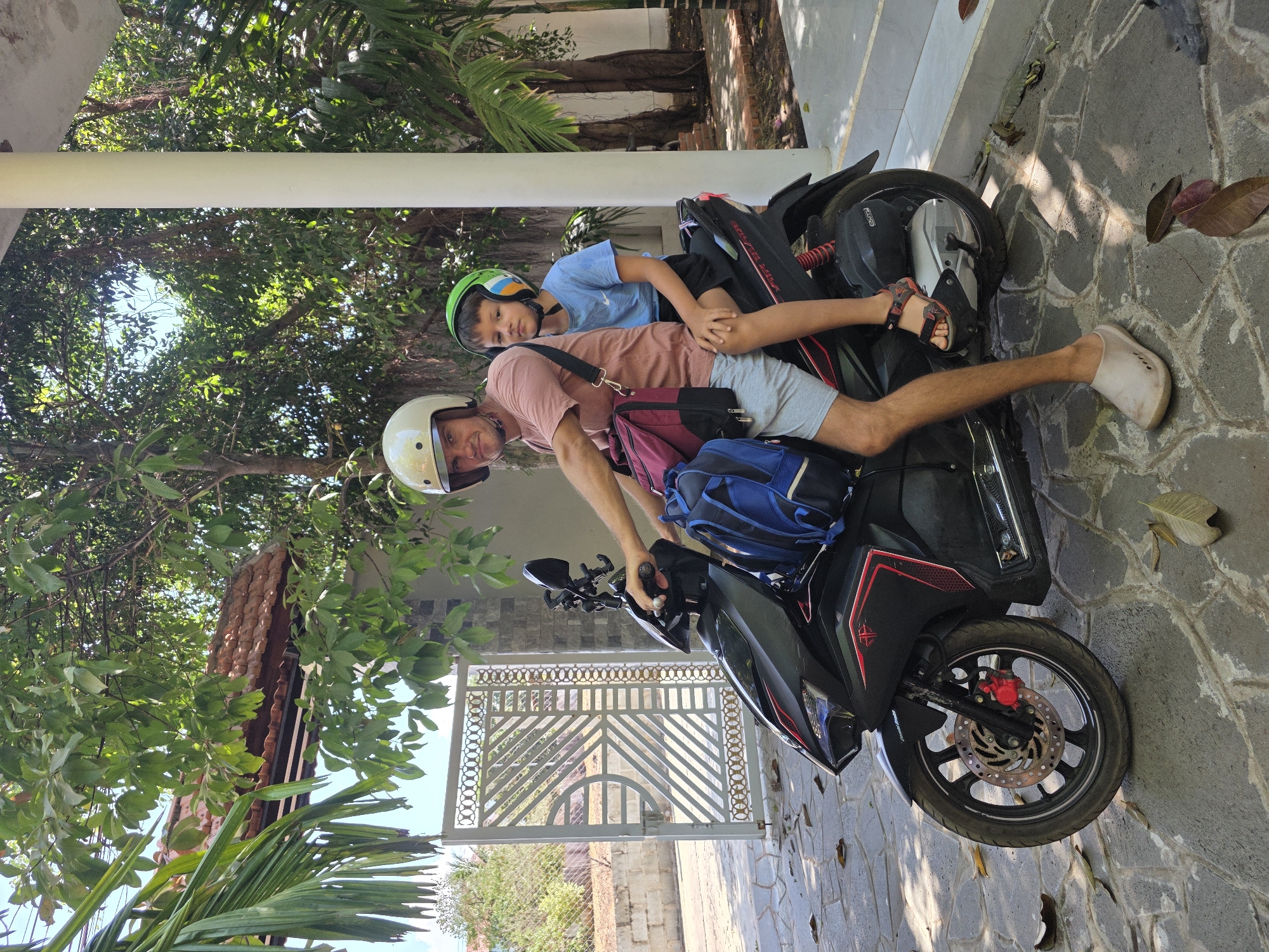 The image shows a man and a child on a black and red scooter in front of a house with a metal gate and lush greenery. The man, wearing a helmet, a light pink t-shirt, and gray shorts, is sitting on the scooter and appears to be preparing to drive. He has a blue backpack and a smaller black and maroon bag slung over his shoulder. The child, wearing a green helmet and a blue shirt, is lying on top of the man. The setting is outdoors, with trees and foliage visible in the background and a house with a tiled roof on the left. The ground is paved with stone, indicating a yard or driveway. The location is inferred to be Mũi Né, Vietnam, based on environmental factors and architectural details typical of the region.