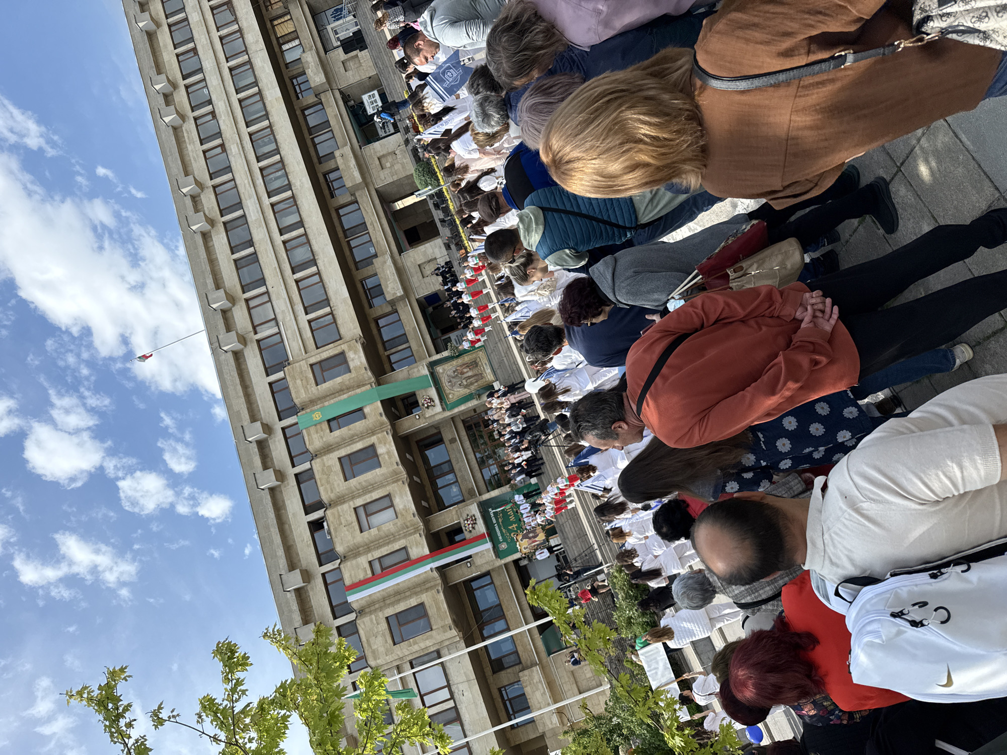 The image shows a crowd of people gathered in front of a large building in Shumen, Bulgaria. The building appears to be a government or administrative building, decorated with Bulgarian flags and banners. Many people are wearing white shirts or dresses. Some people are holding books or notebooks. The crowd appears to be attending a ceremony or event related to education or culture. The sky is blue with some clouds, and there are trees visible in the foreground.