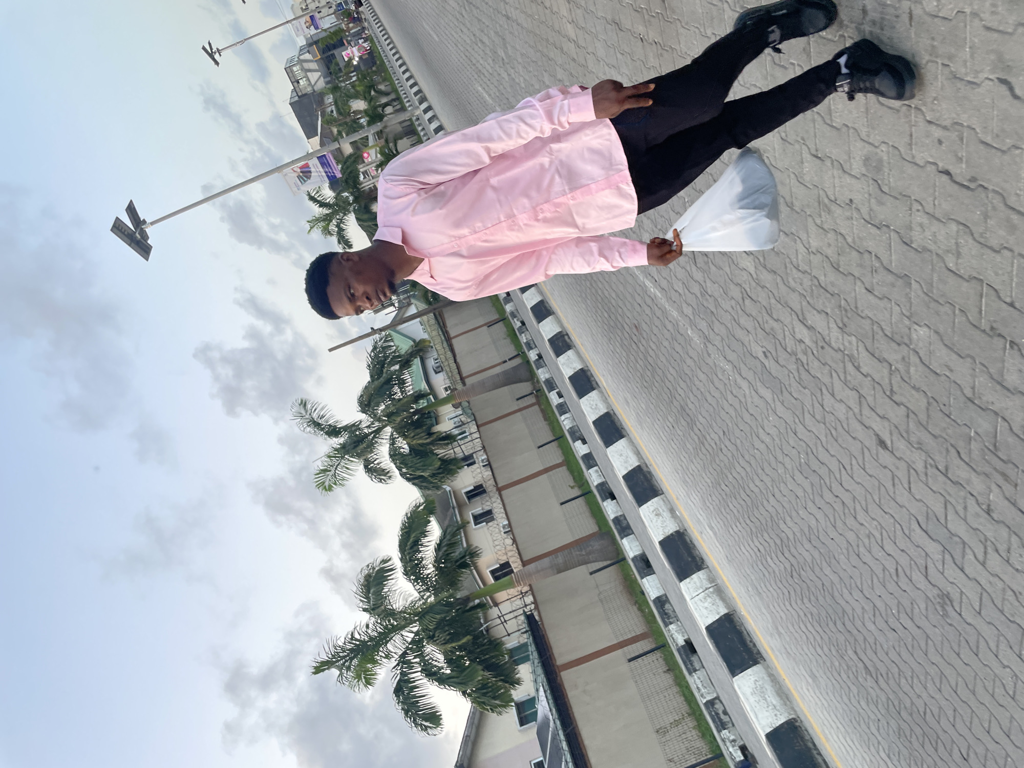A young man in a pink shirt, black pants, and black shoes stands on a paved walkway, holding a white plastic bag. Palm trees line the street and a multi-story building can be seen in the background. The sky is overcast. The location is Ilado, Nigeria, as specified.